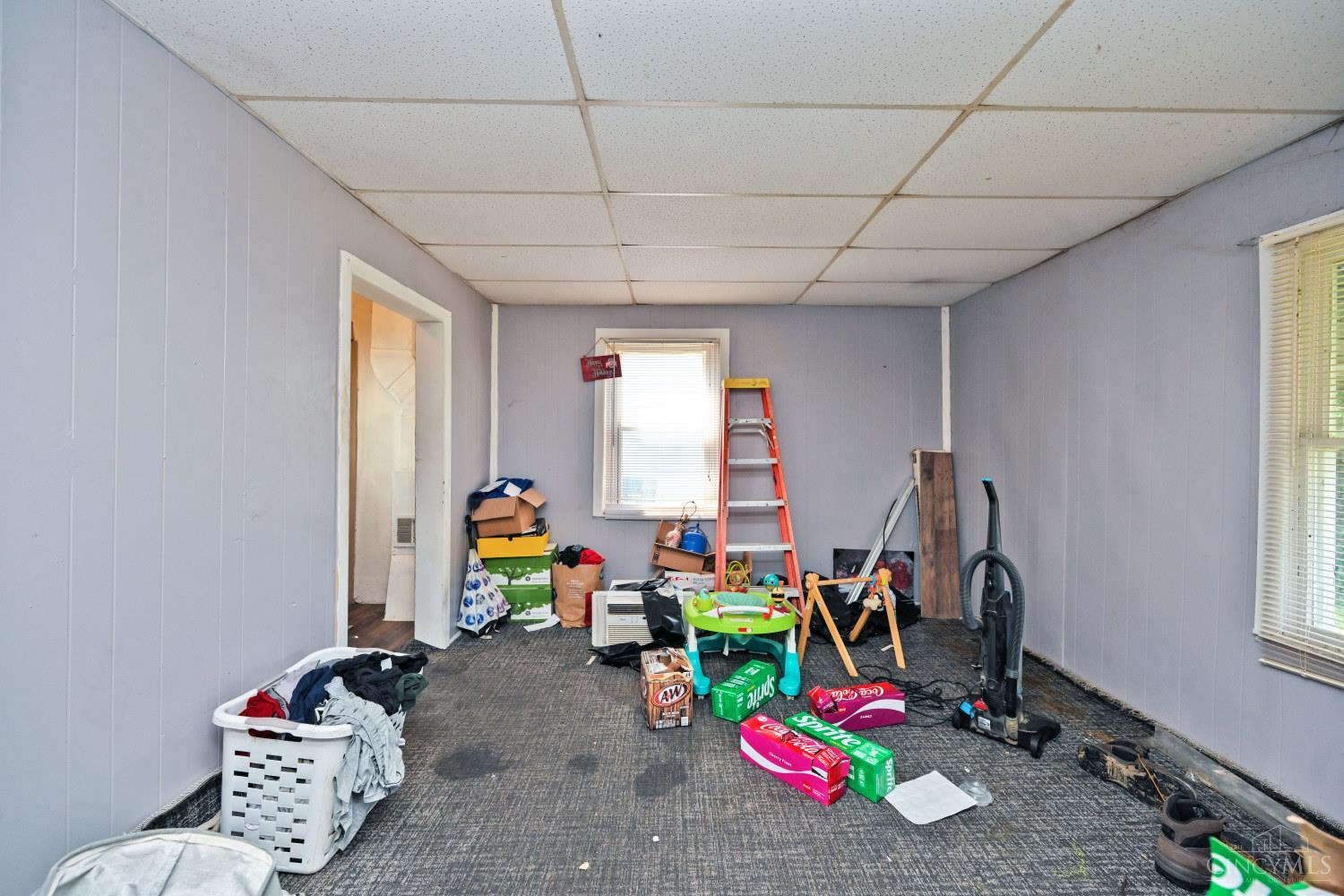 Cluttered carpeted room with a drop ceiling, two windows, a ladder, and stored household items.