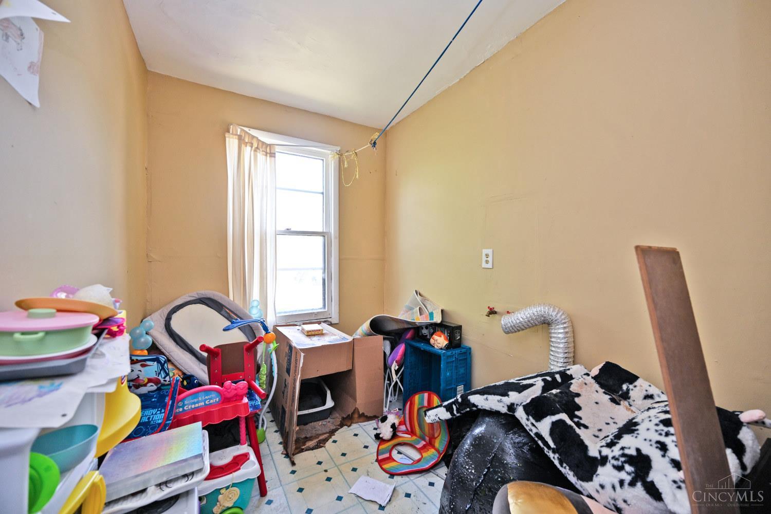 Cluttered bedroom with a curtained window, toys, boxes, and a bed covered in a black-and-white blanket.