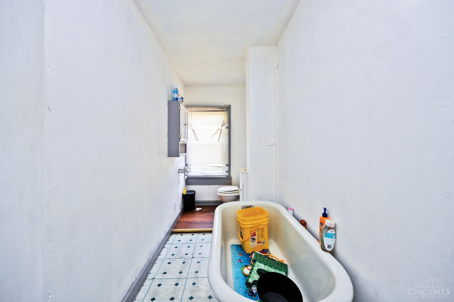 Narrow bathroom with a bathtub full of cleaning supplies, a toilet, and a window with blinds.