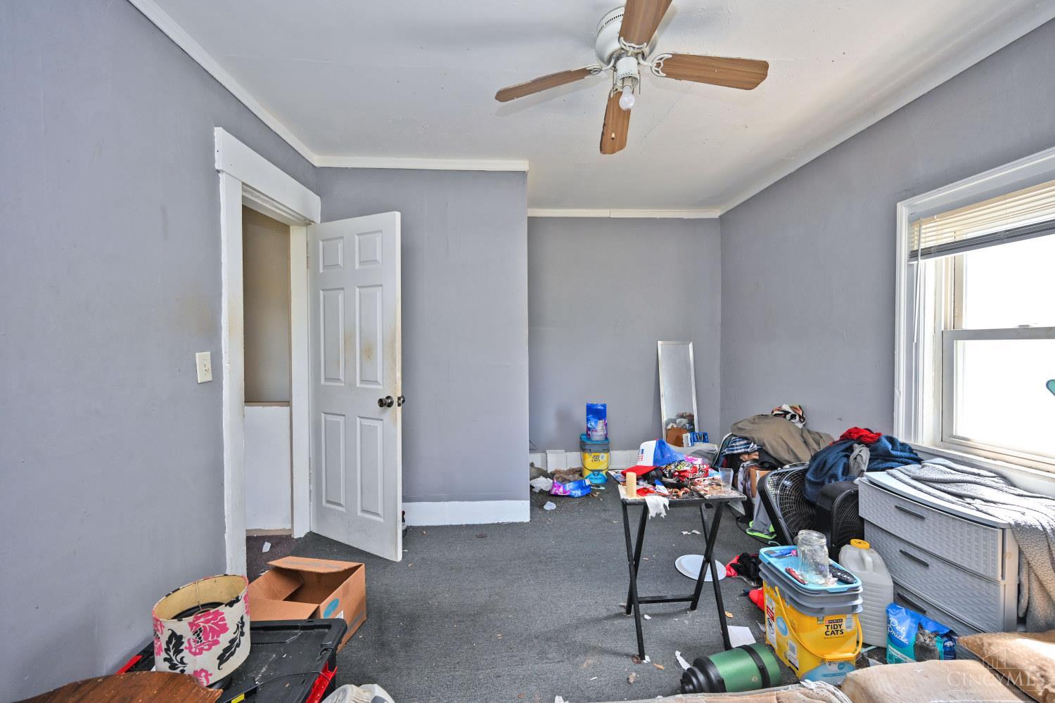Cluttered room with gray walls, dark carpet, a ceiling fan, and a window with blinds.