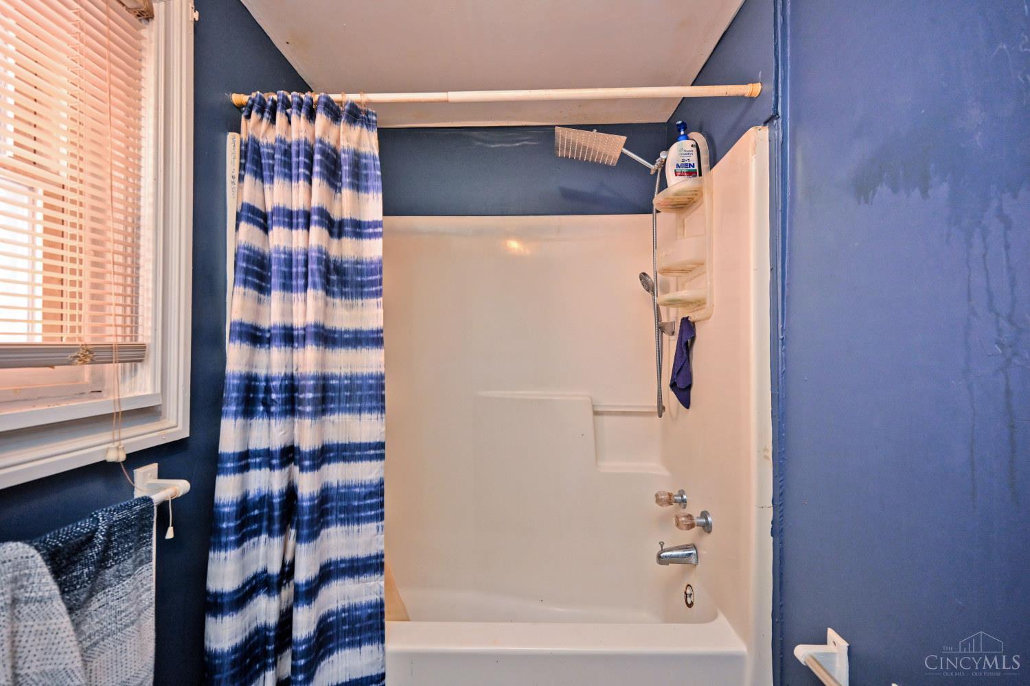 Bathroom with a white tub and shower, blue walls, striped curtain, and window with blinds.