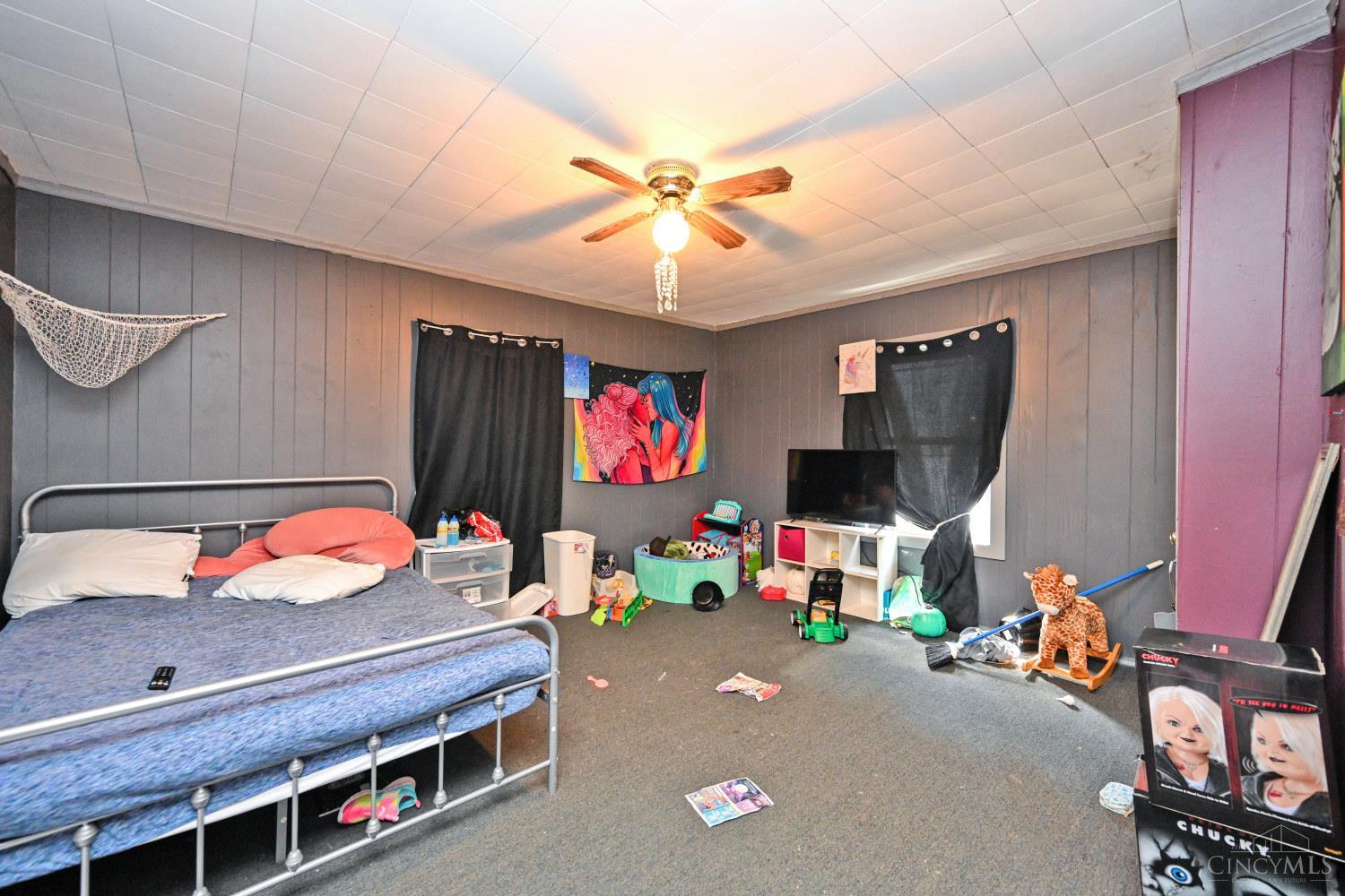 Bedroom with gray paneled walls, a metal bed, ceiling fan, toys, and a television.