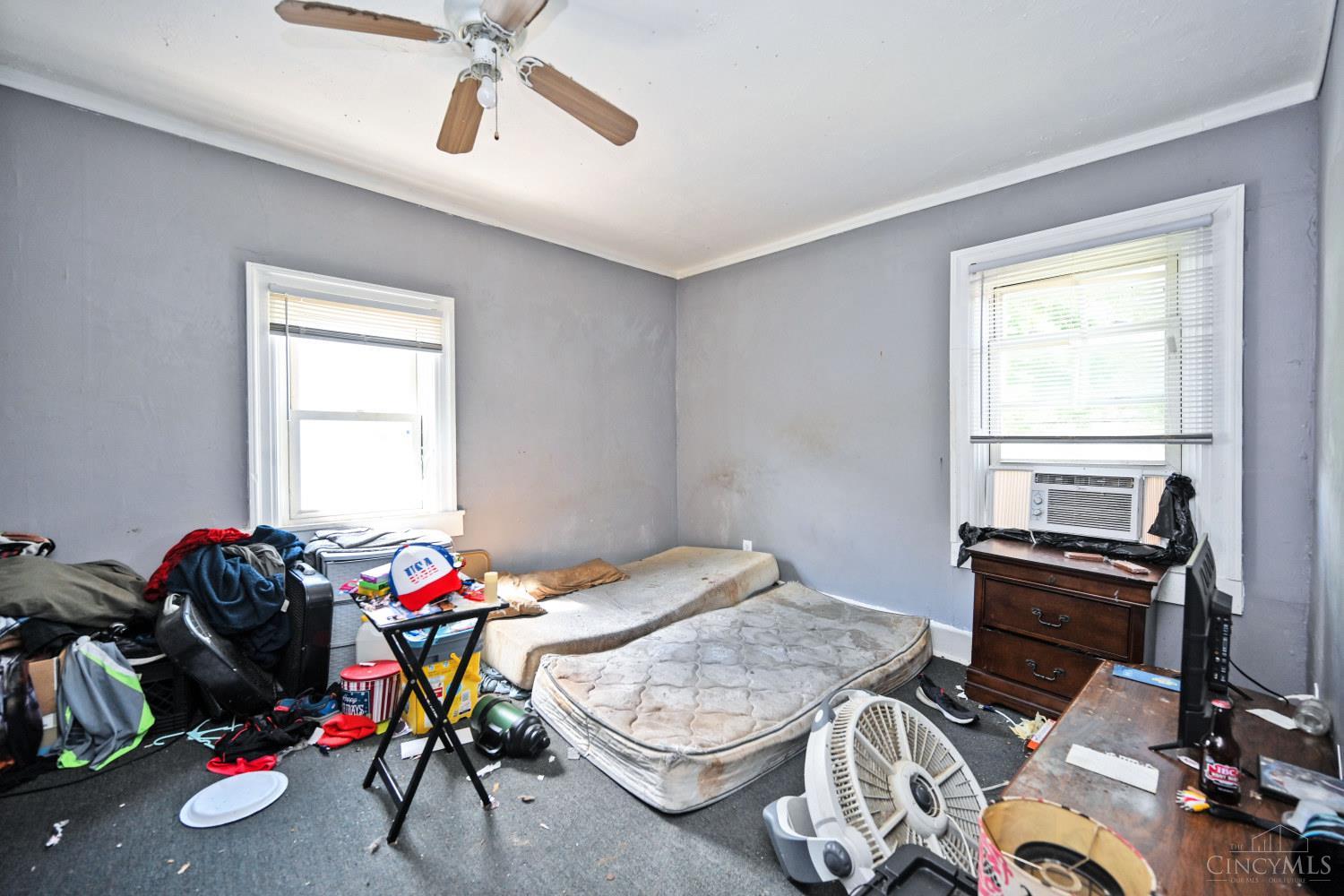 Cluttered bedroom with gray walls, two windows, a ceiling fan, and mattresses on the floor.
