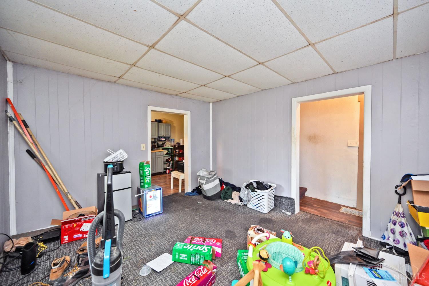 Cluttered basement room with gray paneled walls, a drop ceiling, and doorways to adjoining rooms.