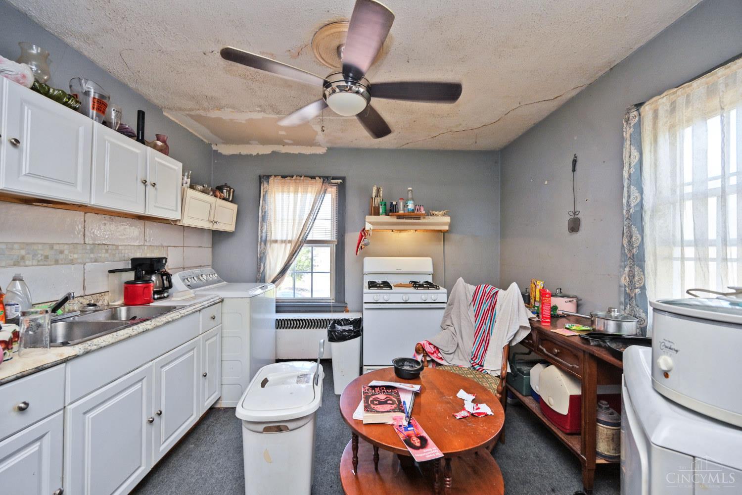 Cluttered kitchen with white cabinets, a gas stove, and a cracked, peeling ceiling.