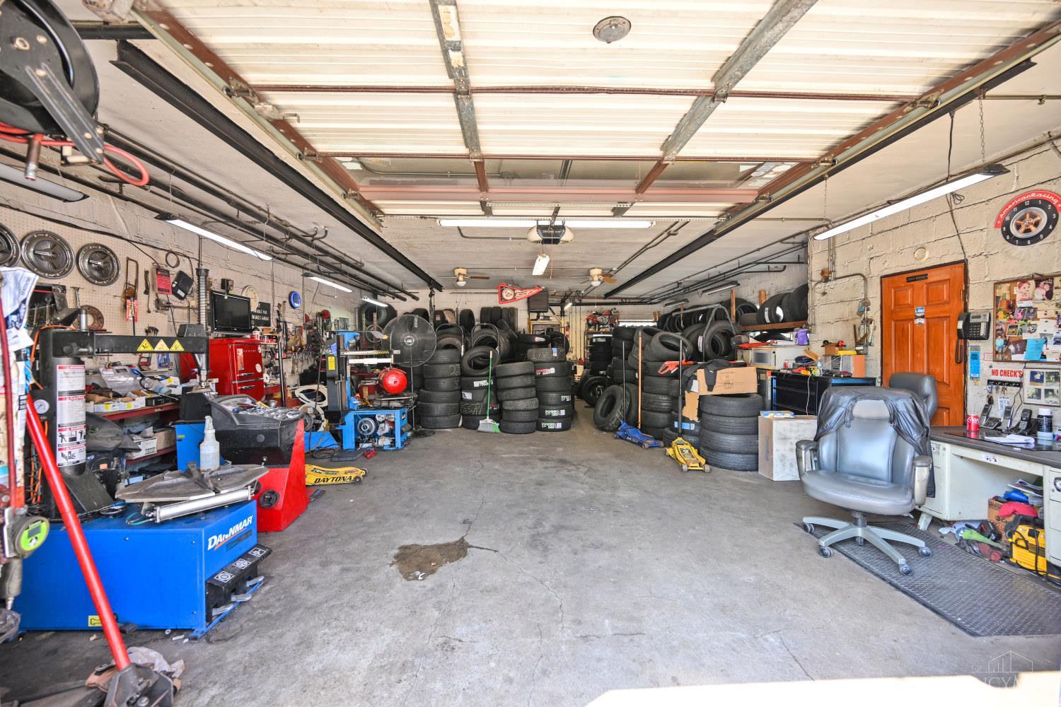 Automotive garage filled with stacked tires, repair equipment, and workbenches beneath an open overhead door.