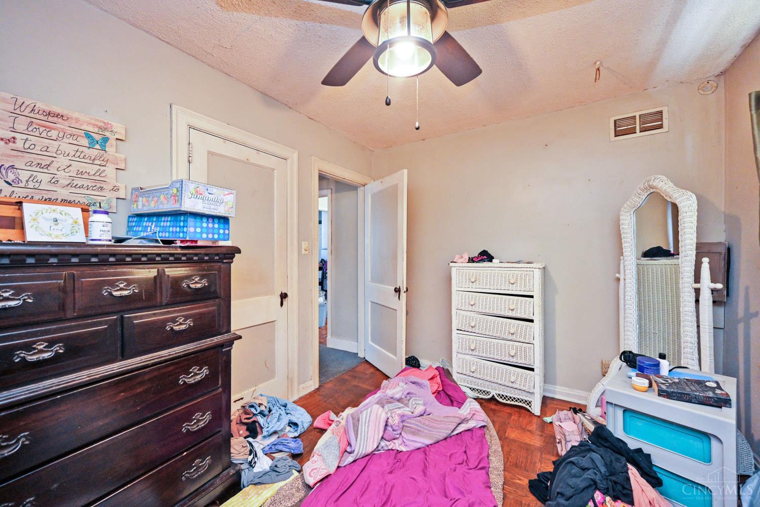 Cluttered bedroom with a dark dresser, white wicker furniture, and clothes and bedding scattered on the floor.