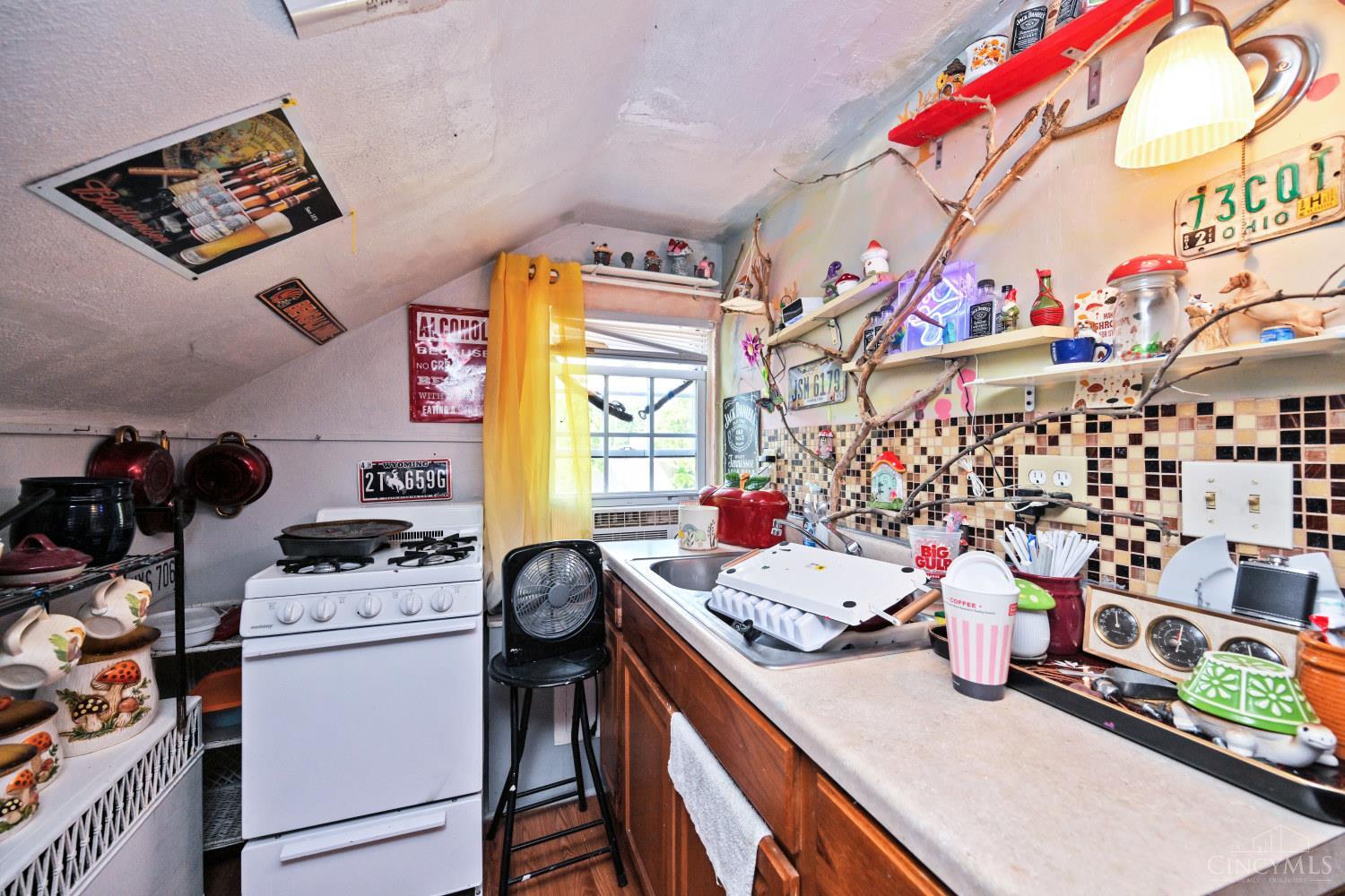 Compact kitchen with a white stove, crowded counters, tiled backsplash, and a window above the sink.