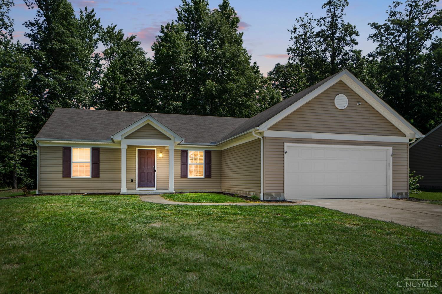 Single-story beige home with a two-car garage, covered entry, and lawn at dusk.