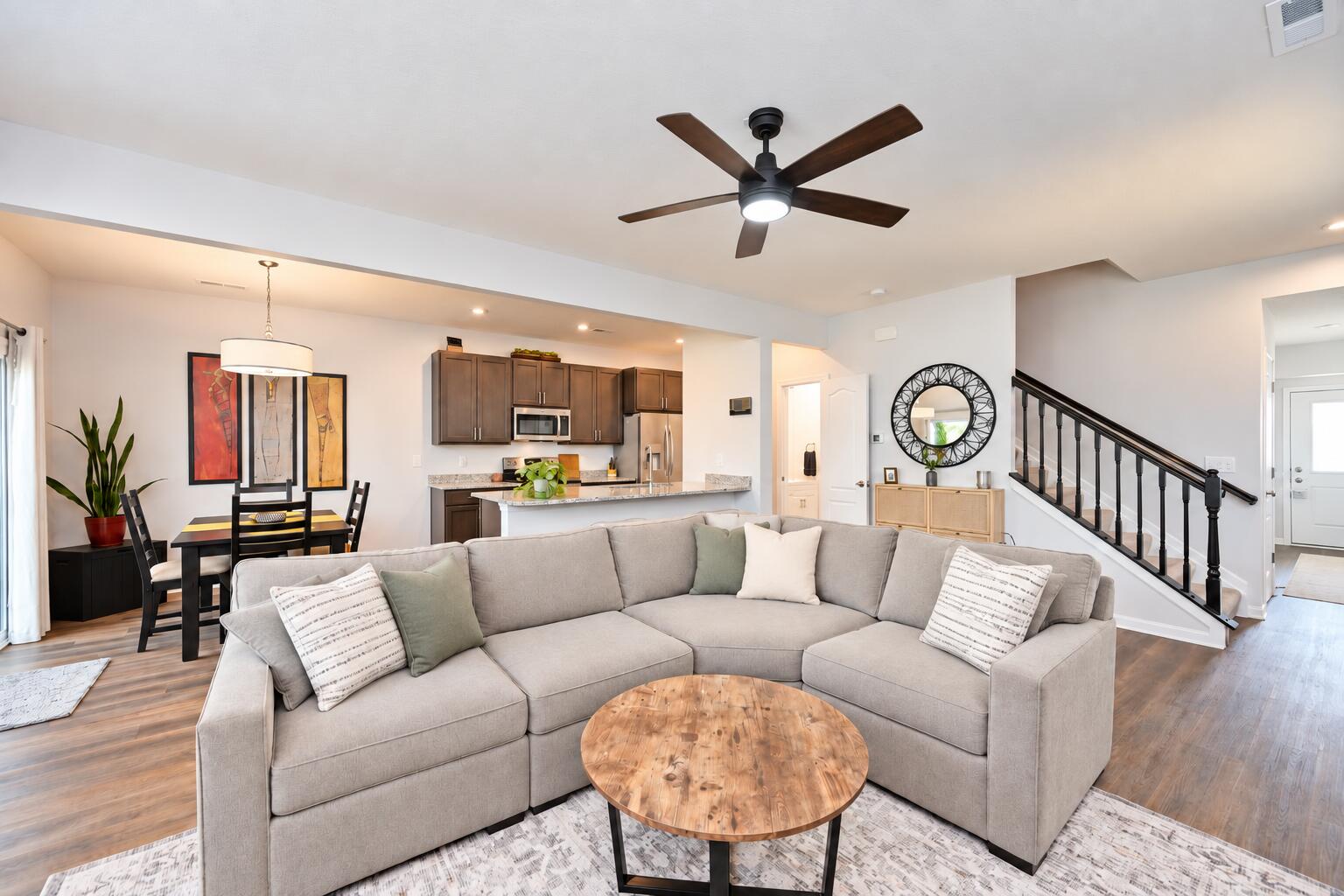 Open-plan living room with a gray sectional, wood coffee table, dining area, and dark-cabinet kitchen.