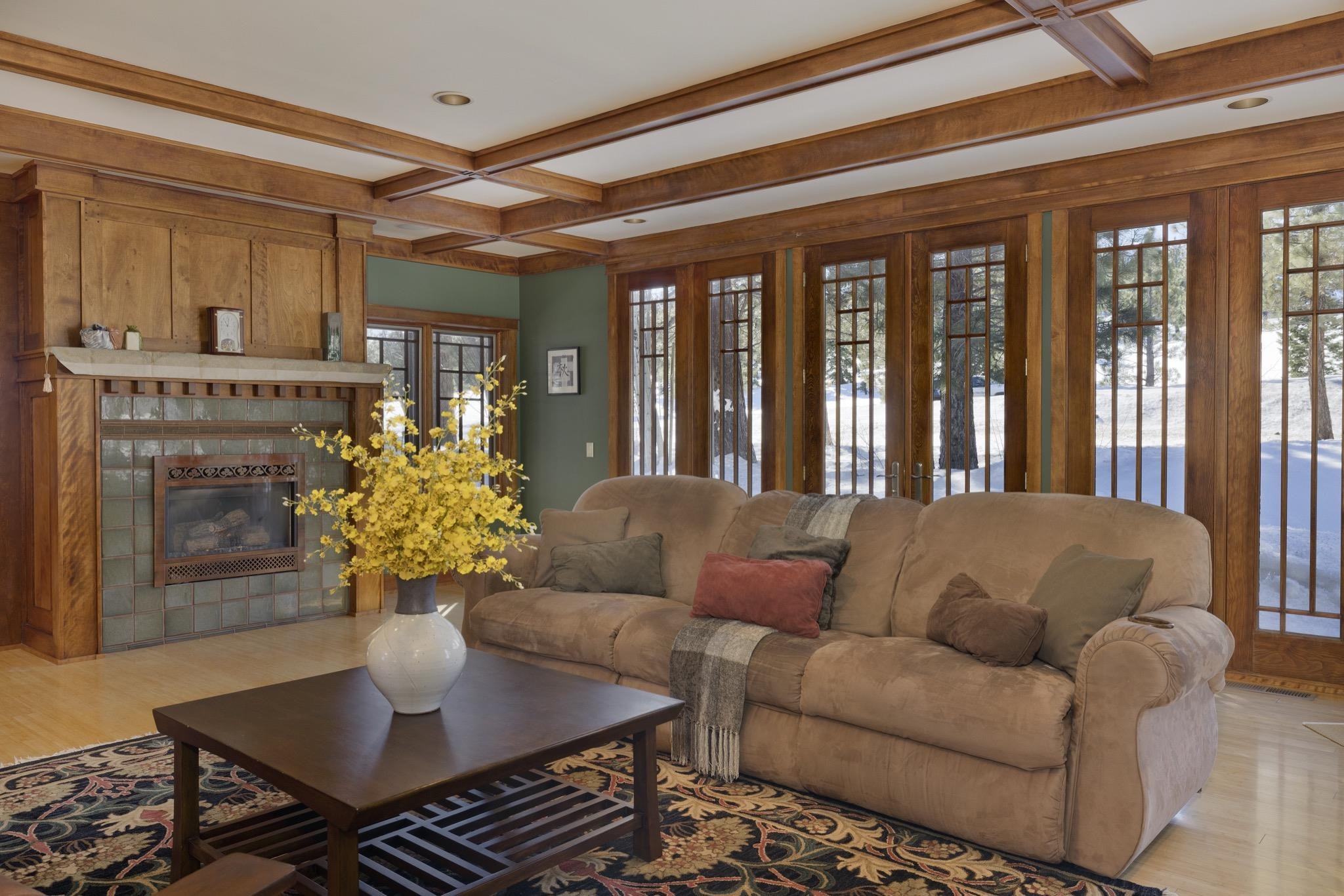 Living room with a tiled fireplace, wood ceiling beams, and tall windows overlooking snowy trees.