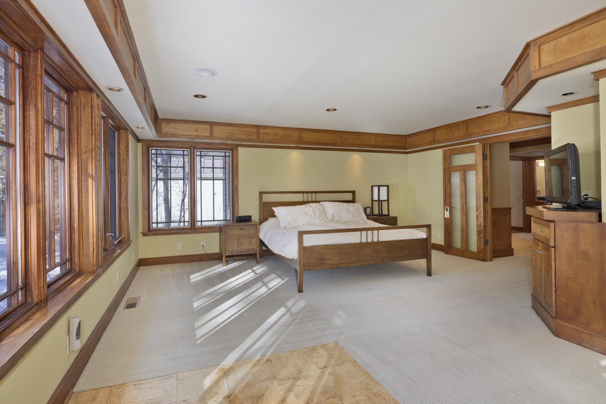 Bedroom with a wooden bed and large wood-trimmed windows casting sunlight across the carpet.