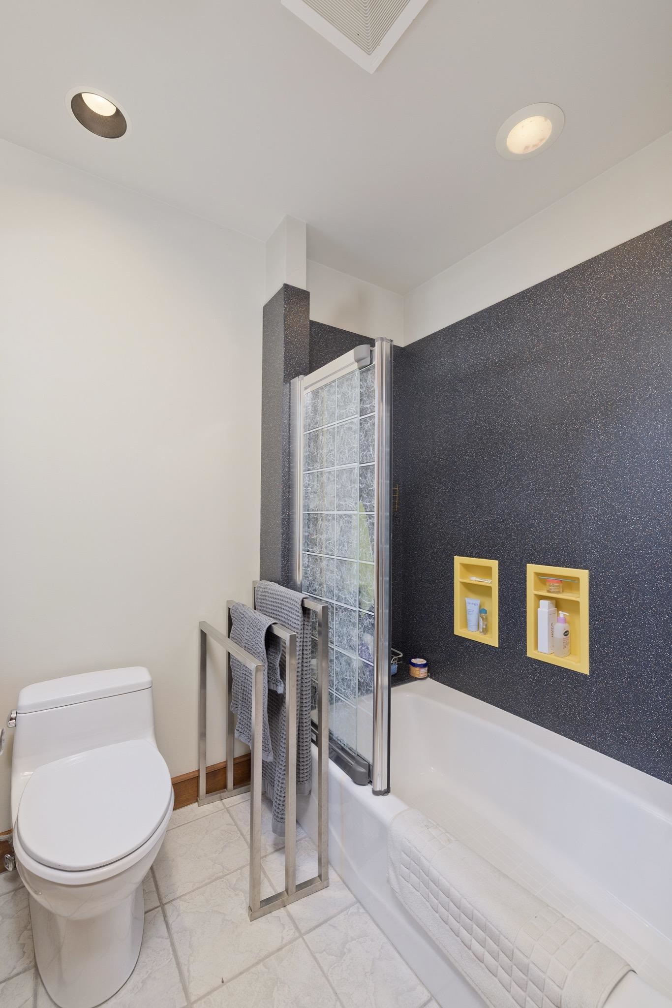 Bathroom with a bathtub, glass-block shower partition, dark accent wall, and yellow recessed shelves.