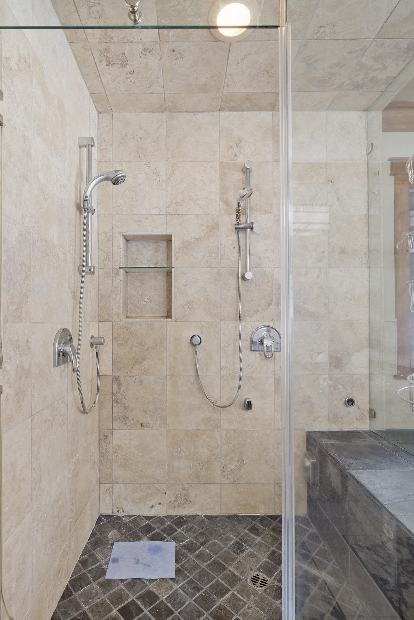 Walk-in shower with dual handheld showerheads, a recessed shelf, and dark tile flooring.