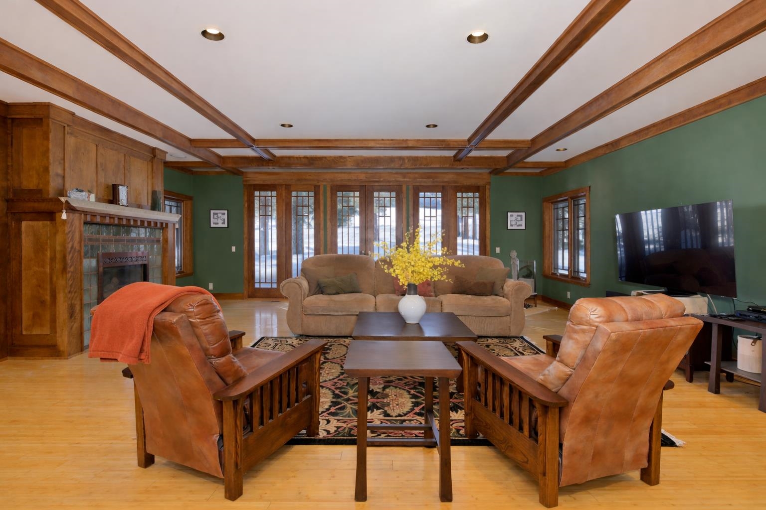 Living room with a stone fireplace, wood ceiling beams, leather chairs, and tall windows overlooking snow.