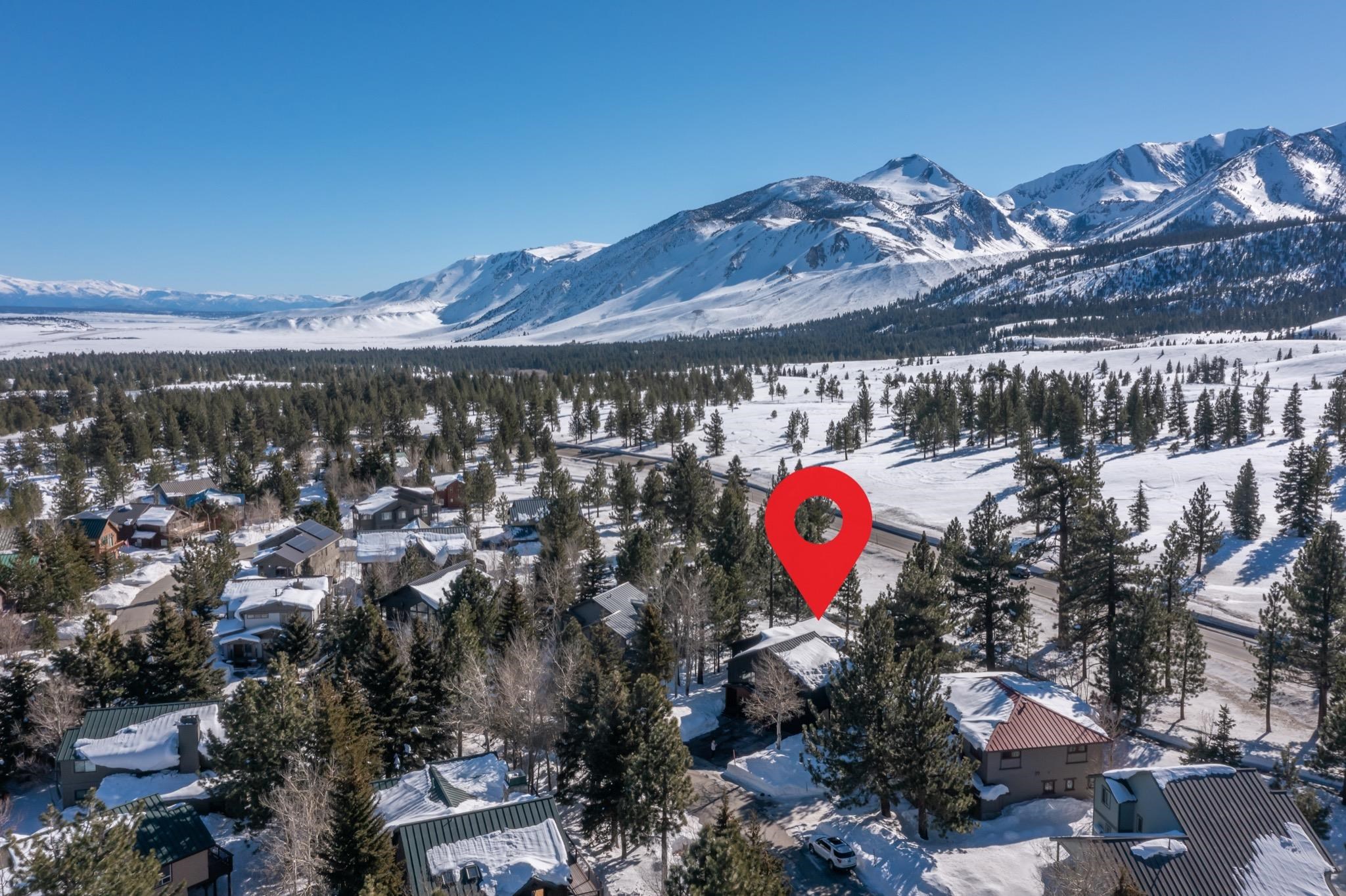 Aerial view of a snow-covered neighborhood beneath mountain peaks, with a red marker over one home.