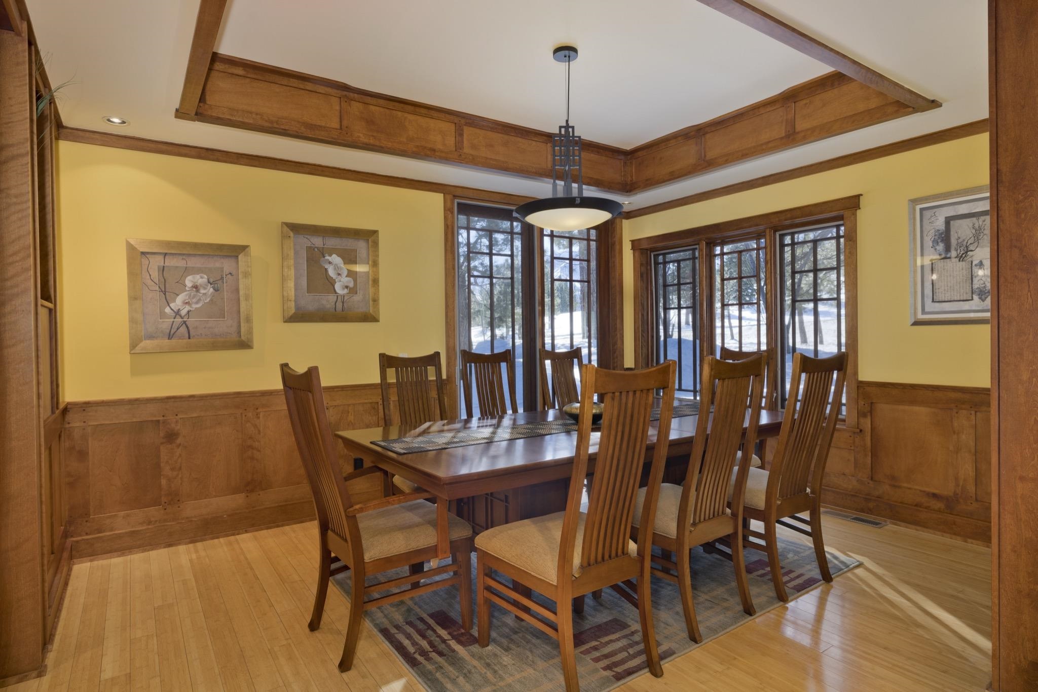 Dining room with a wooden table and chairs, wood-paneled walls, and large windows overlooking snowy trees.