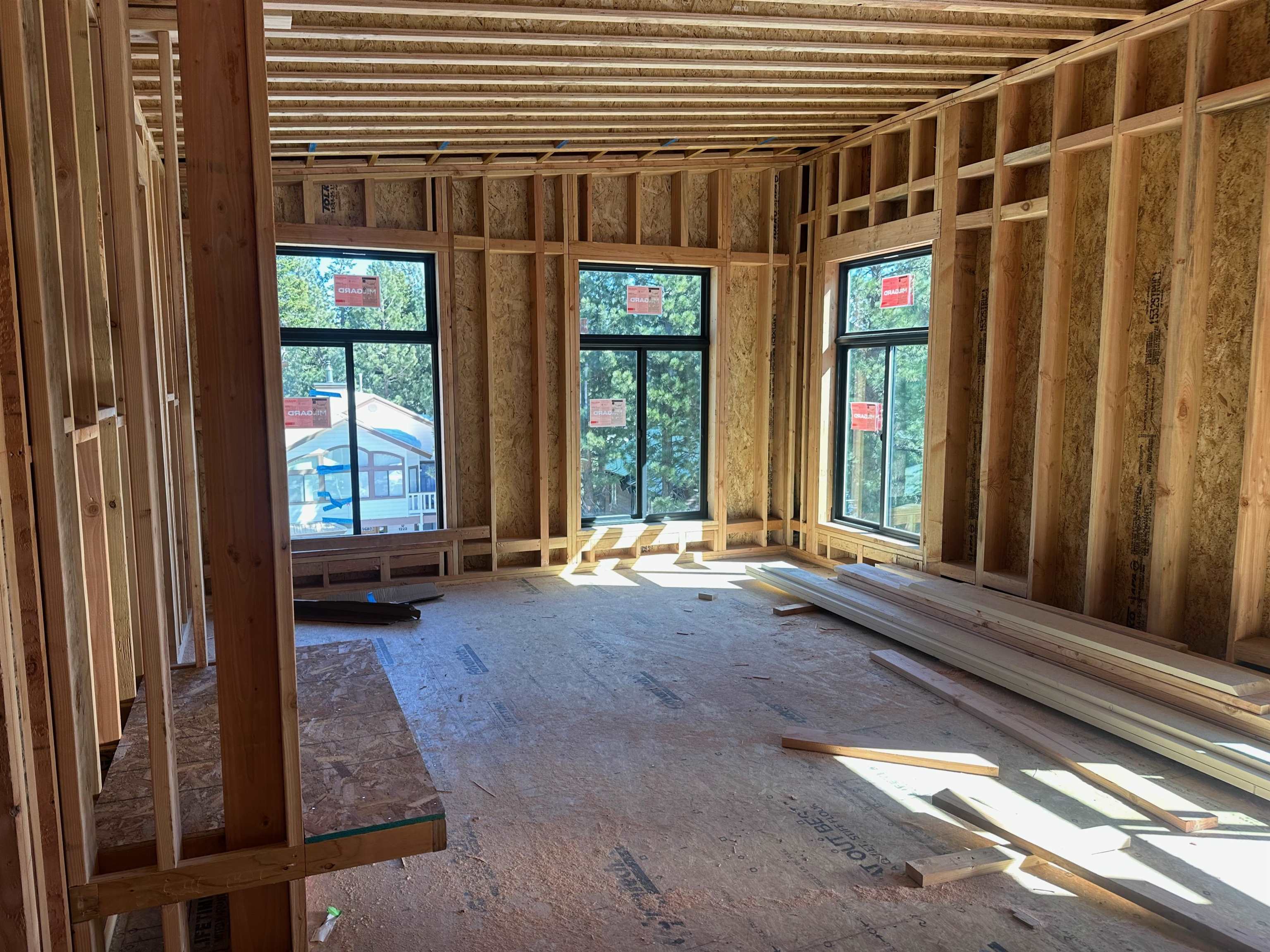 Unfinished framed room with three large windows and stacks of lumber on the bare floor.