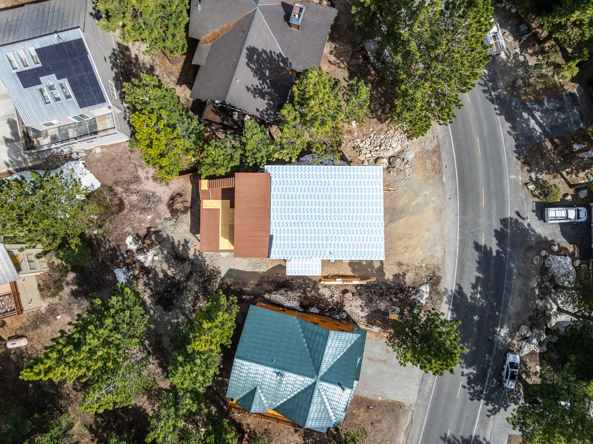 House under construction with exposed roof sheathing, viewed from above among pine trees.