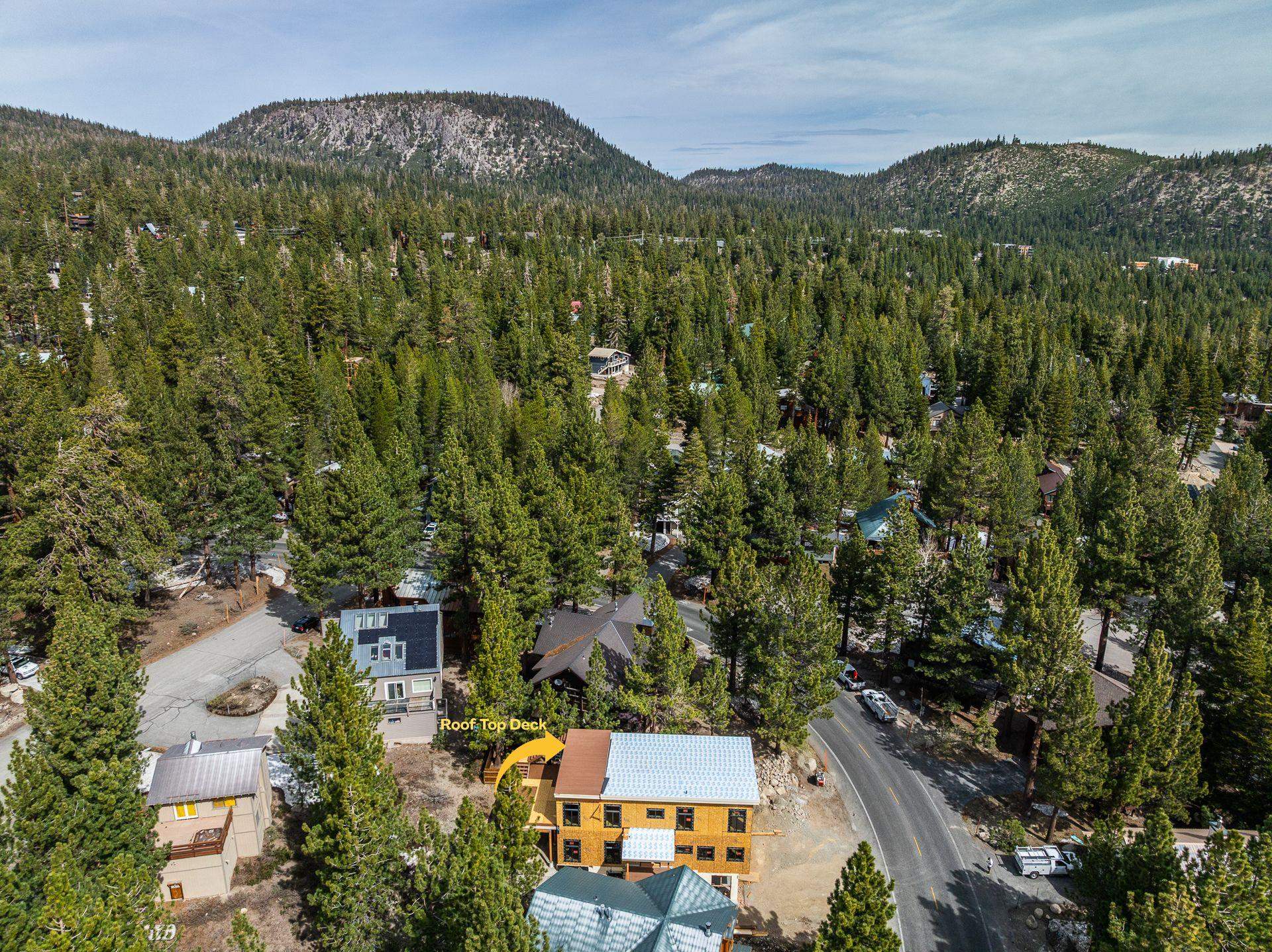 Aerial view of a house under construction, surrounded by pine trees with mountain ridges beyond.