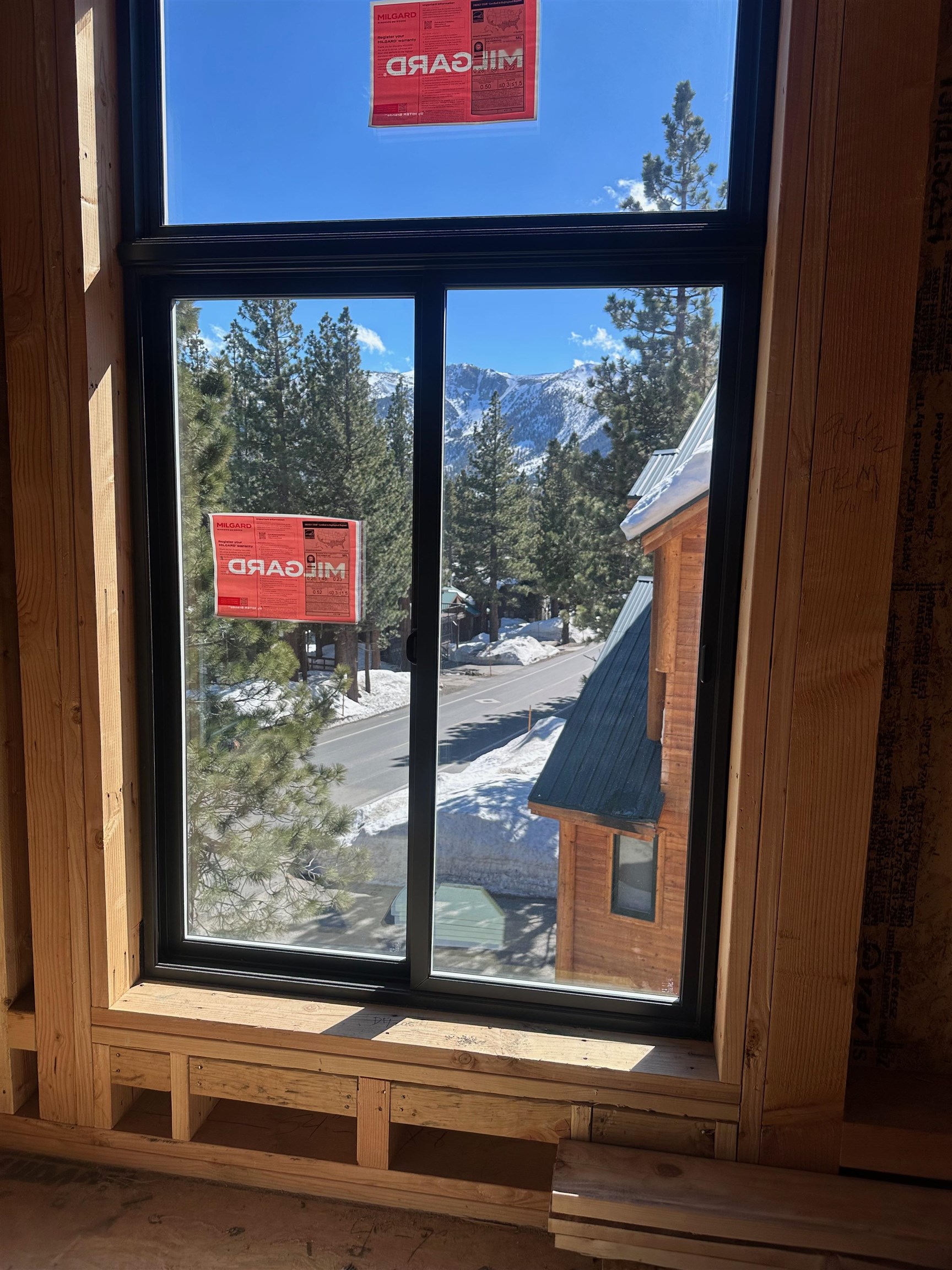 Large window in an unfinished wood interior overlooking snowy mountains and pine trees.
