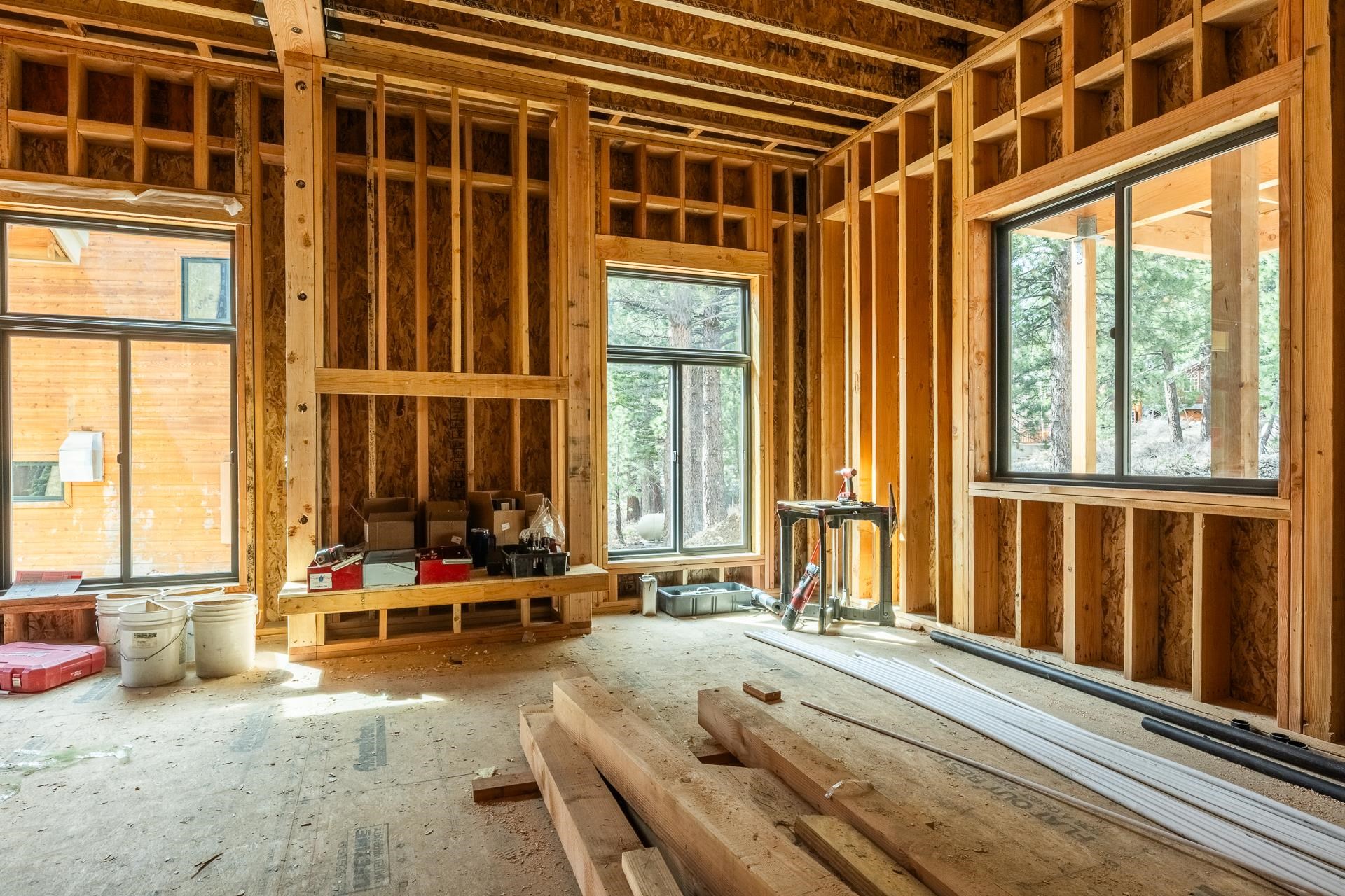 Unfinished room with exposed wood framing, construction materials, and large windows overlooking trees.
