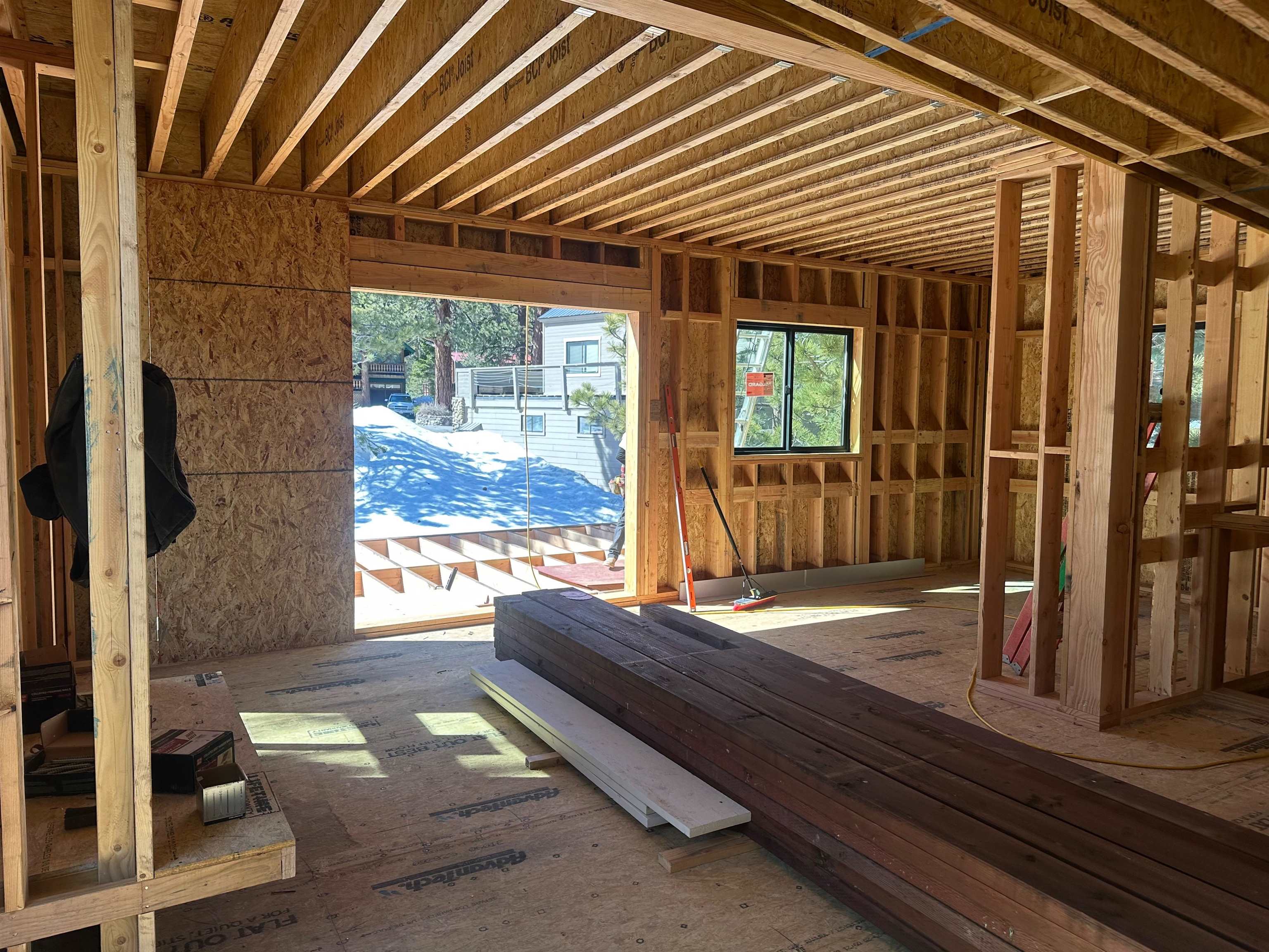 Unfinished house interior with exposed framing, stacked lumber, and snow visible through a large opening.