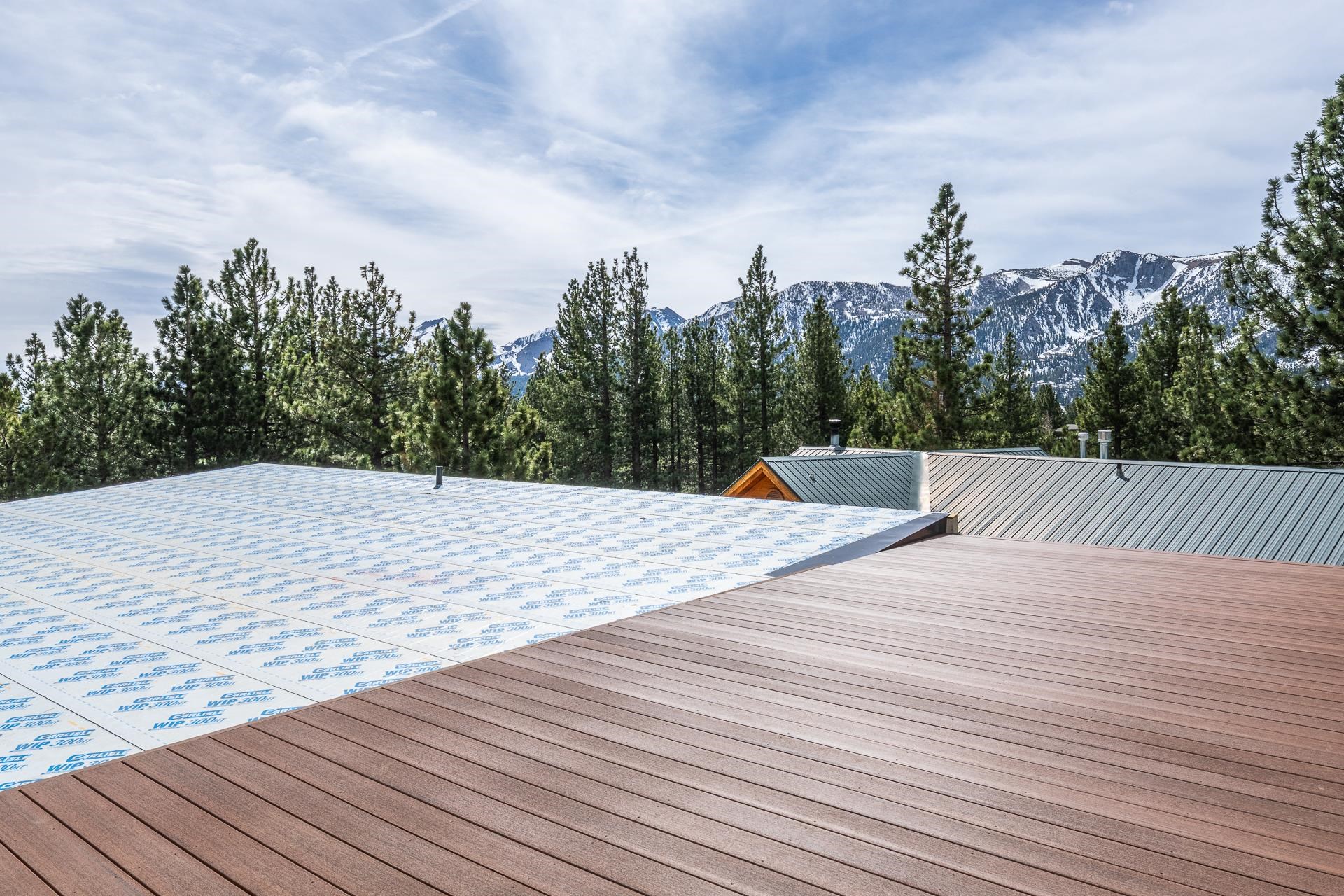 Broad deck overlooking pine forest and snow-dusted mountains, beside a roof with exposed printed underlayment.