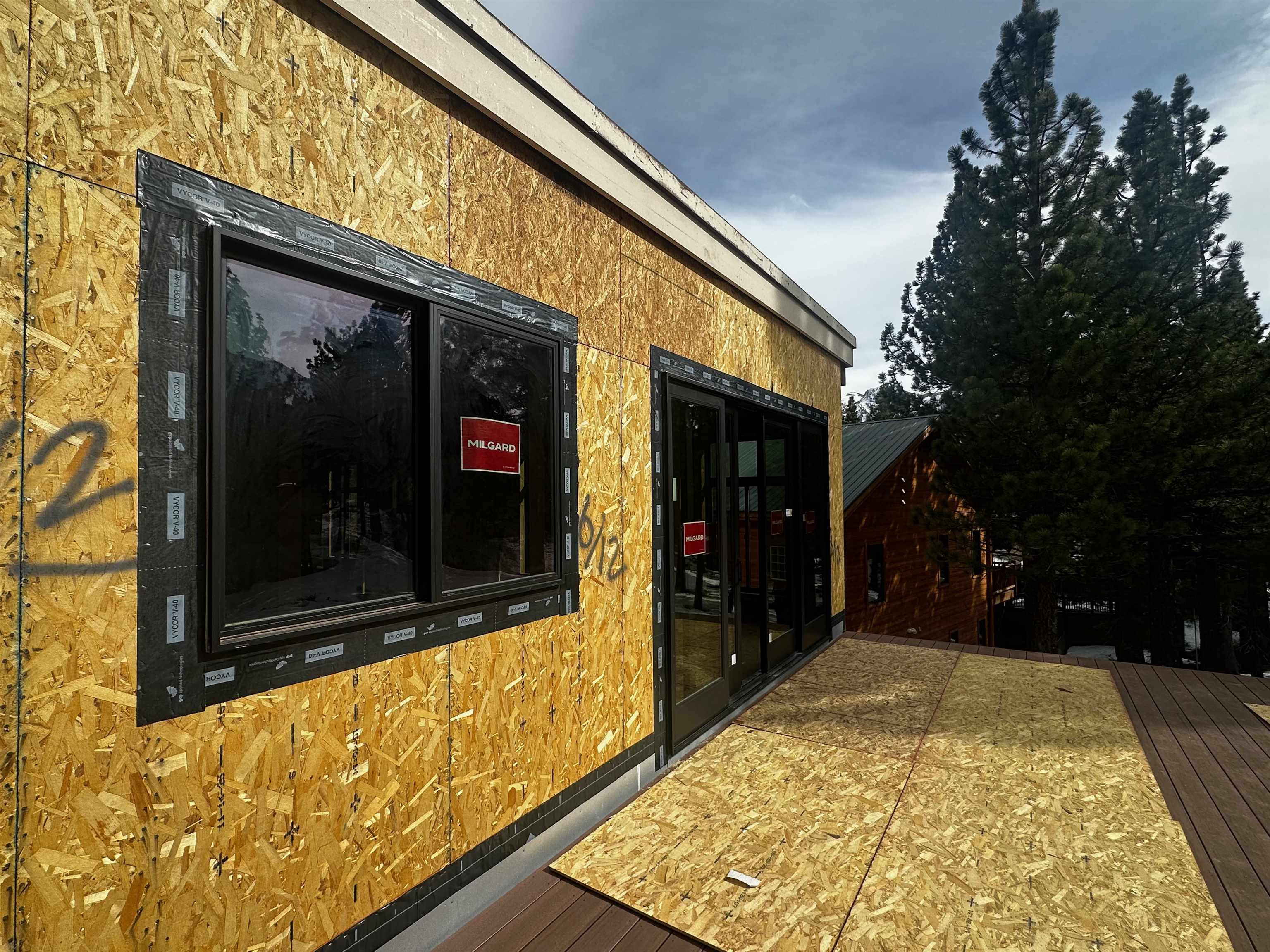 House under construction with exposed plywood sheathing, black-framed windows, and an unfinished deck among pine trees.