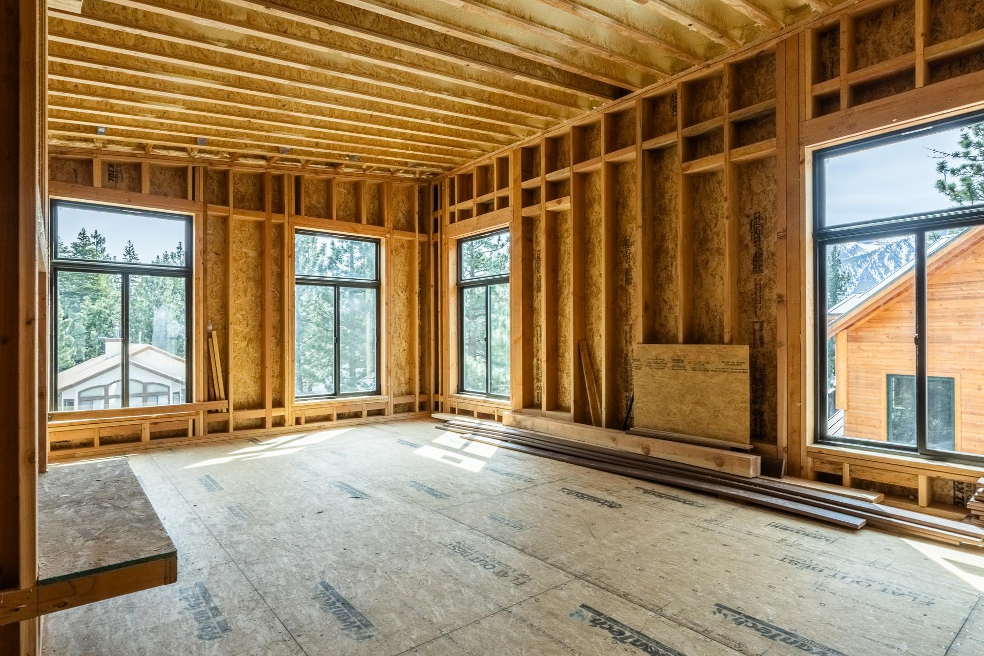 Unfinished interior with exposed framing and large windows overlooking pine trees and a mountain.