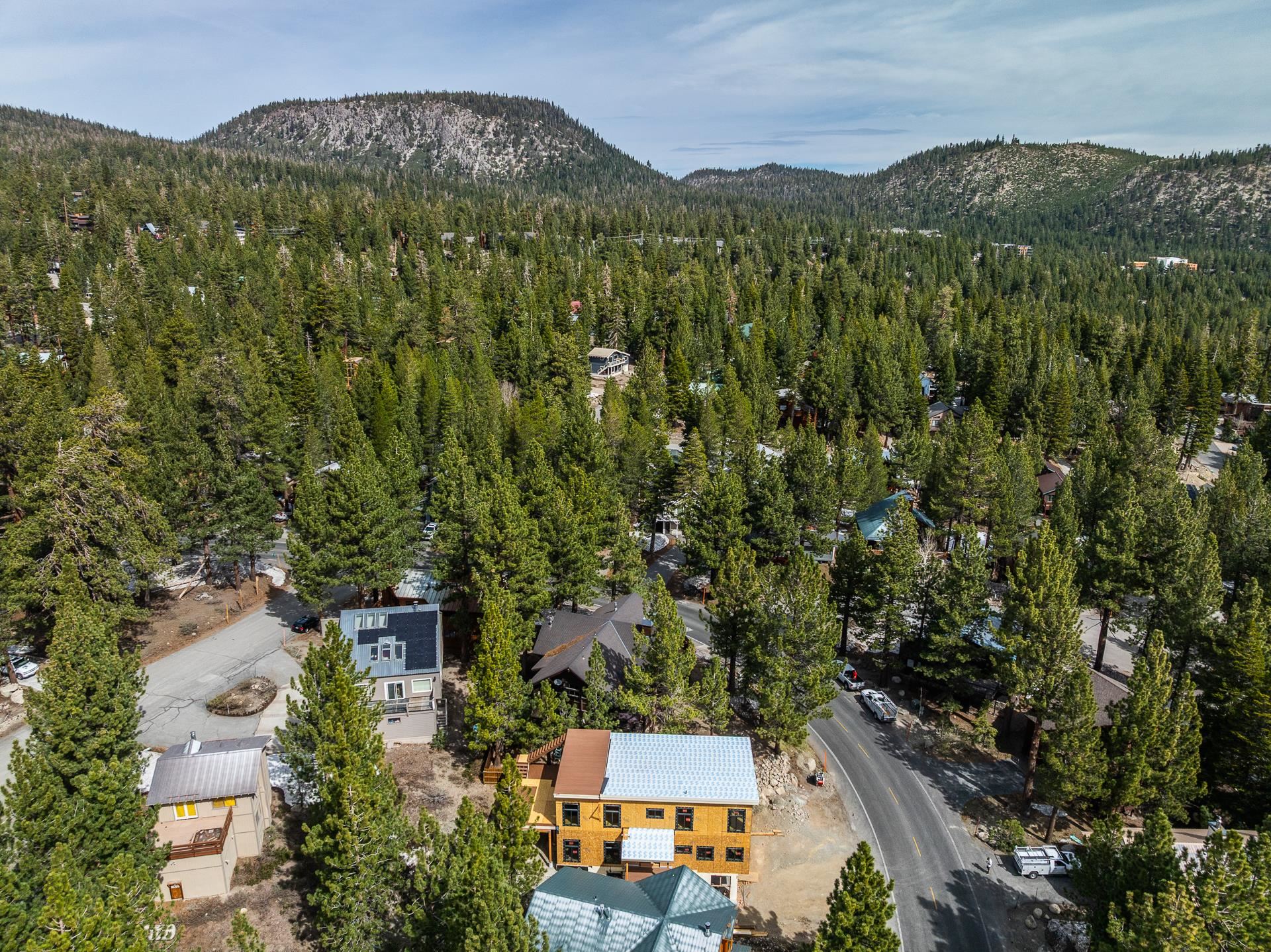 House under construction among pine trees, with a neighborhood road and forested mountains beyond.