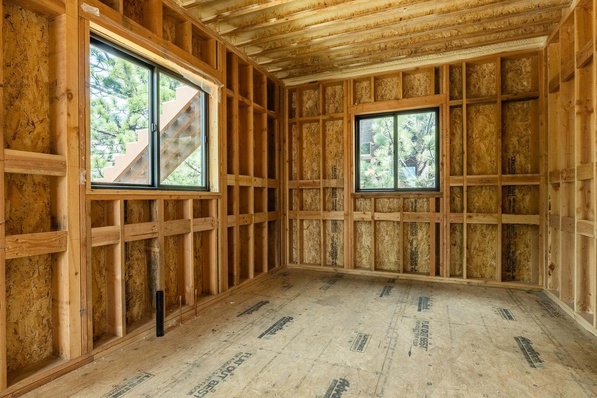 Unfinished room with exposed framing, plywood subfloor, and windows facing pine trees.