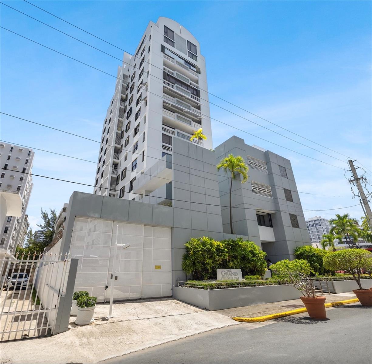 White and gray condominium tower with balconies, a gated entrance, and palm trees.
