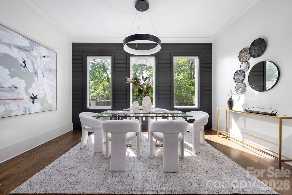 Dining room with a glass table, white upholstered chairs, and a black paneled wall with three windows.