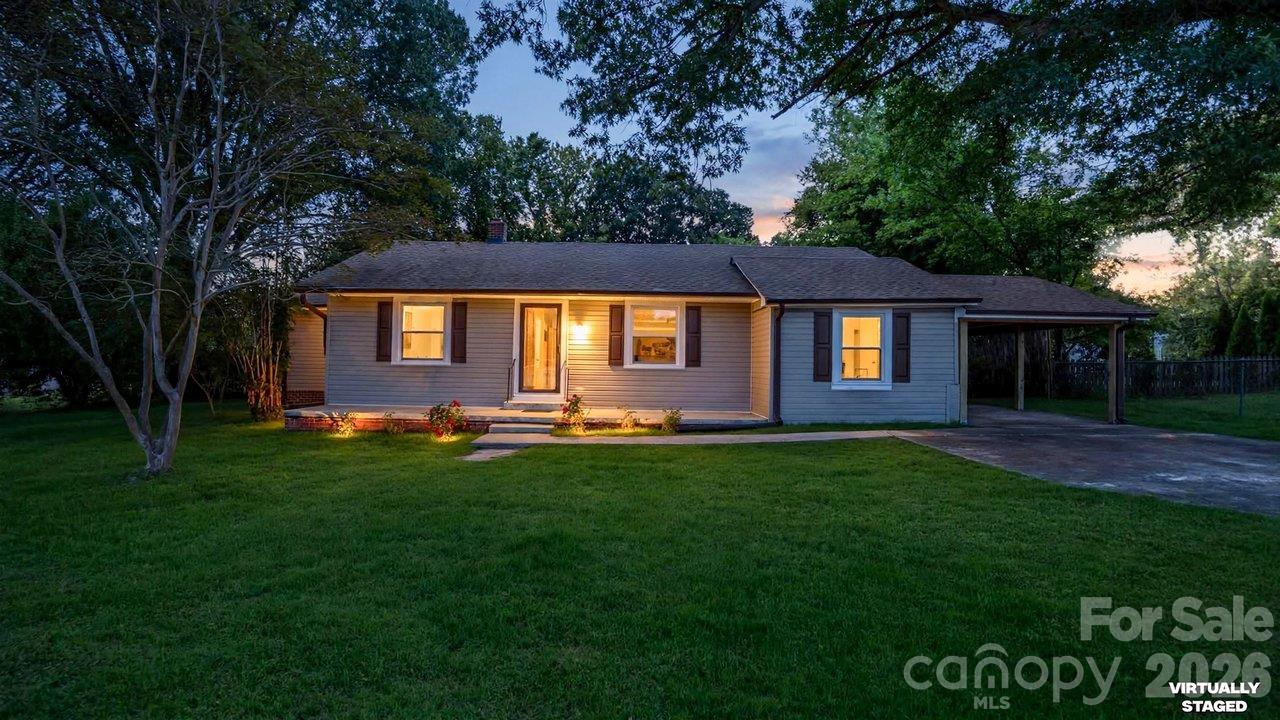 Single-story gray house at dusk, with warmly lit windows, a broad lawn, and an attached carport.