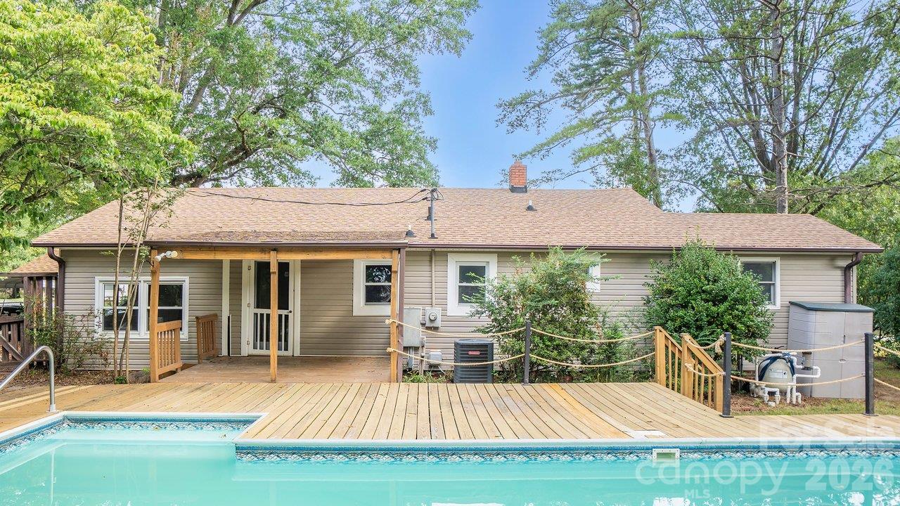 Rear of a gray-sided home with a broad wooden deck and swimming pool beneath mature trees.