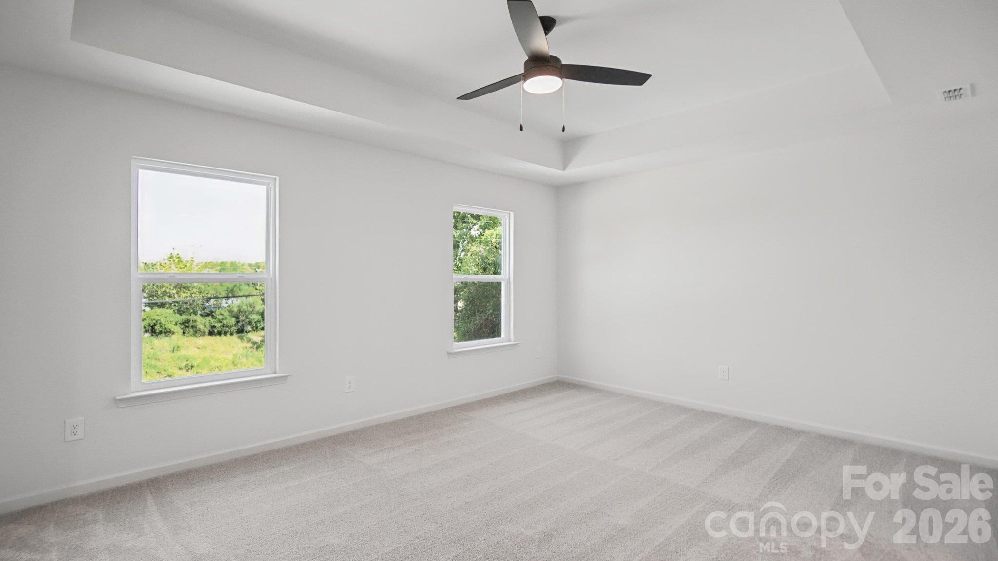 Empty bedroom with gray carpet, two windows overlooking greenery, and a ceiling fan.