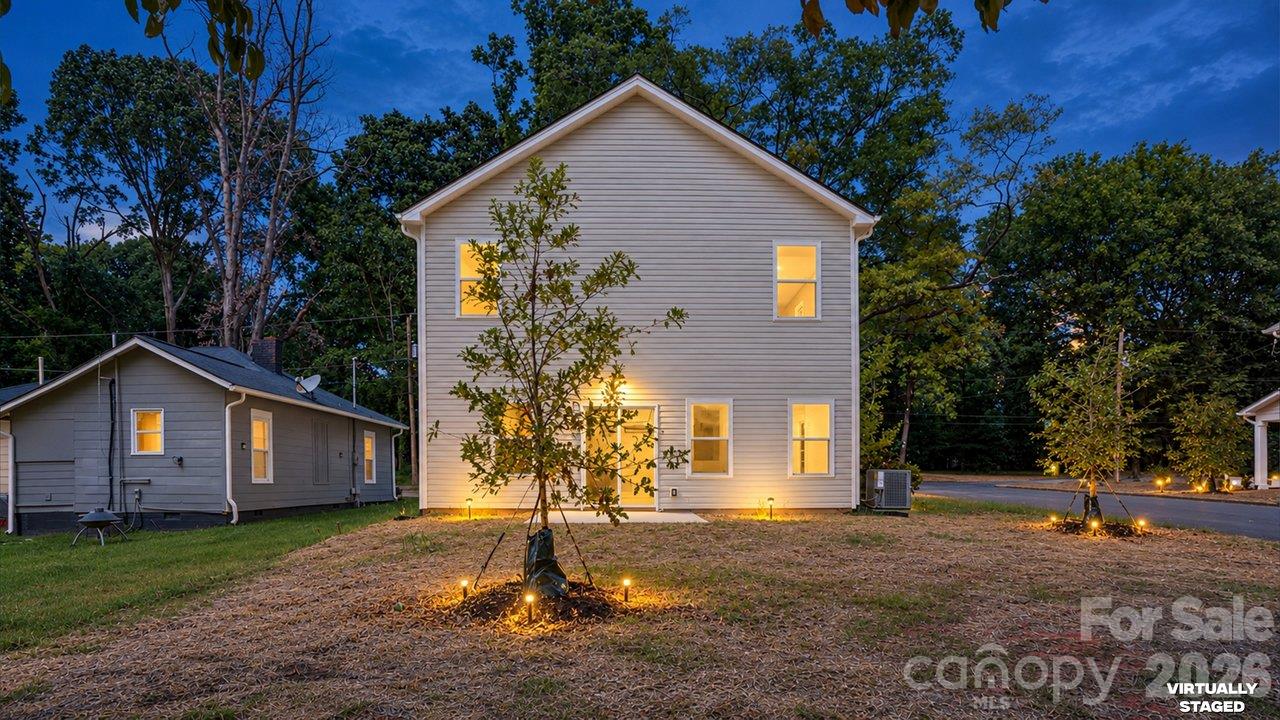 Two-story light-sided house at dusk, with warmly lit windows and young trees in the yard.