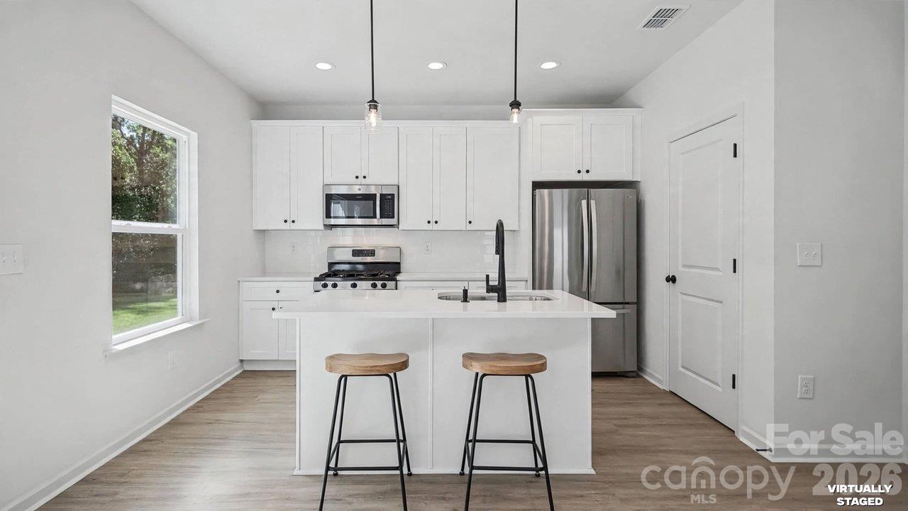 Bright white kitchen with a central island, two stools, stainless steel appliances, and a window overlooking greenery.