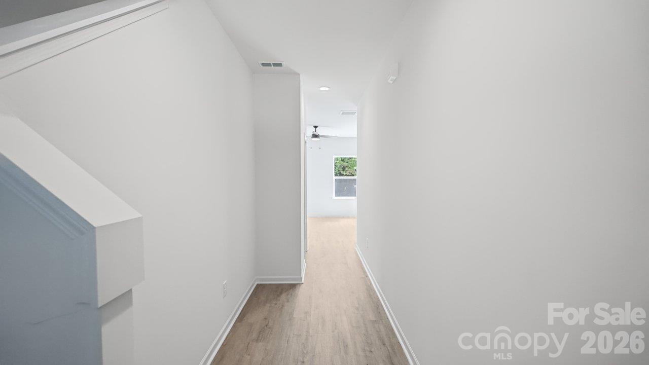 Empty hallway with light wood floors, white walls, and a staircase railing in the foreground.