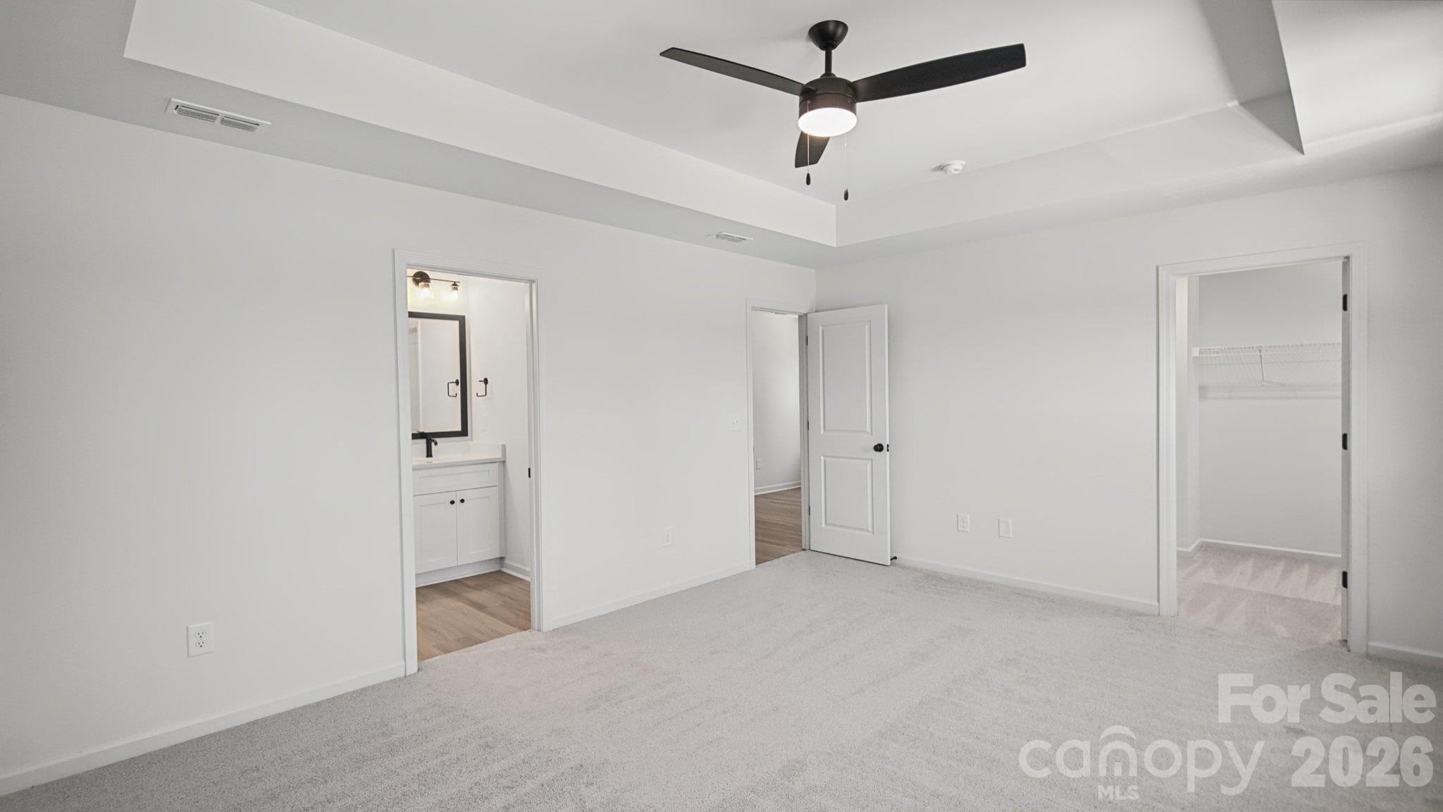 Empty carpeted bedroom with a tray ceiling, black ceiling fan, ensuite bathroom, and closet.