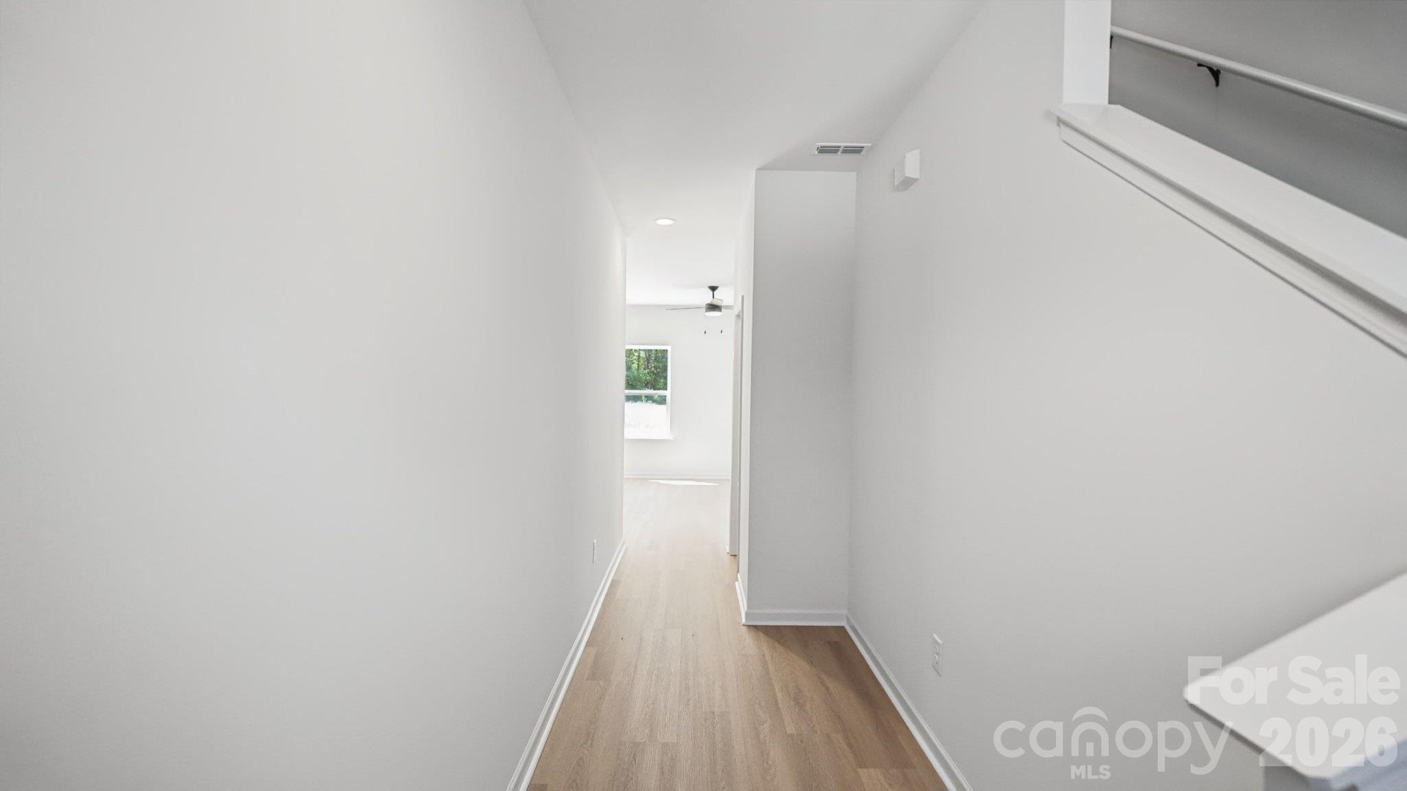 Empty hallway with light wood flooring and a staircase railing leading toward a bright room.