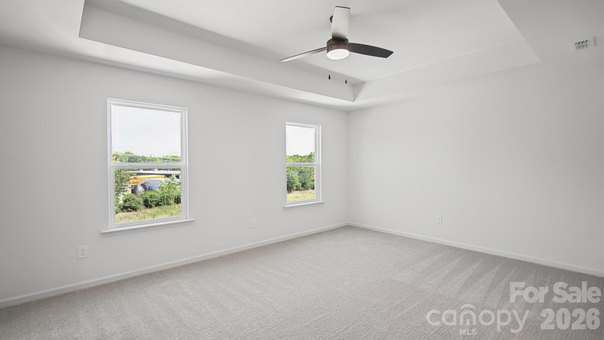 Empty bedroom with gray carpet, two windows, and a ceiling fan beneath a tray ceiling.