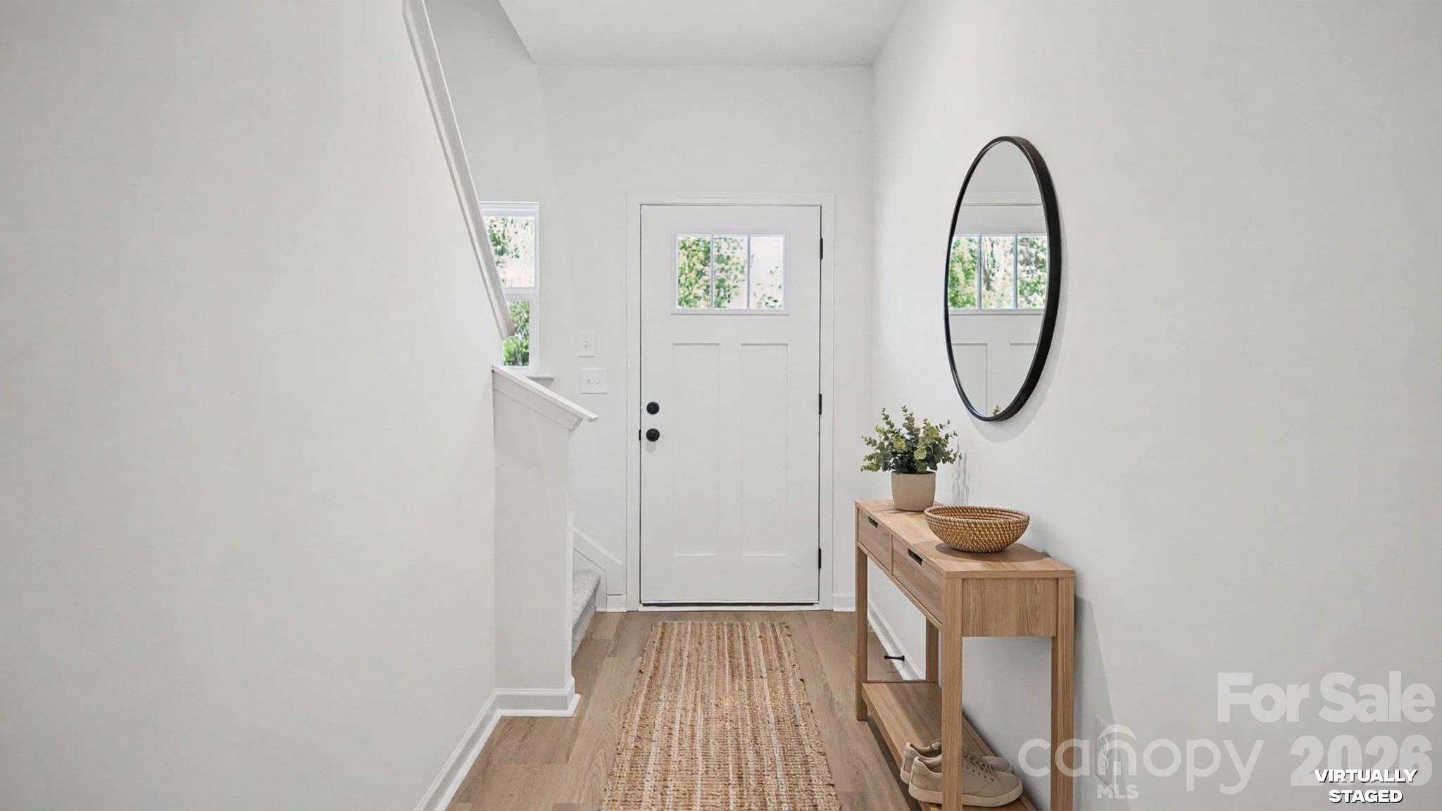 Bright entryway with a white front door, wood floors, woven runner, and console table beneath an oval mirror.