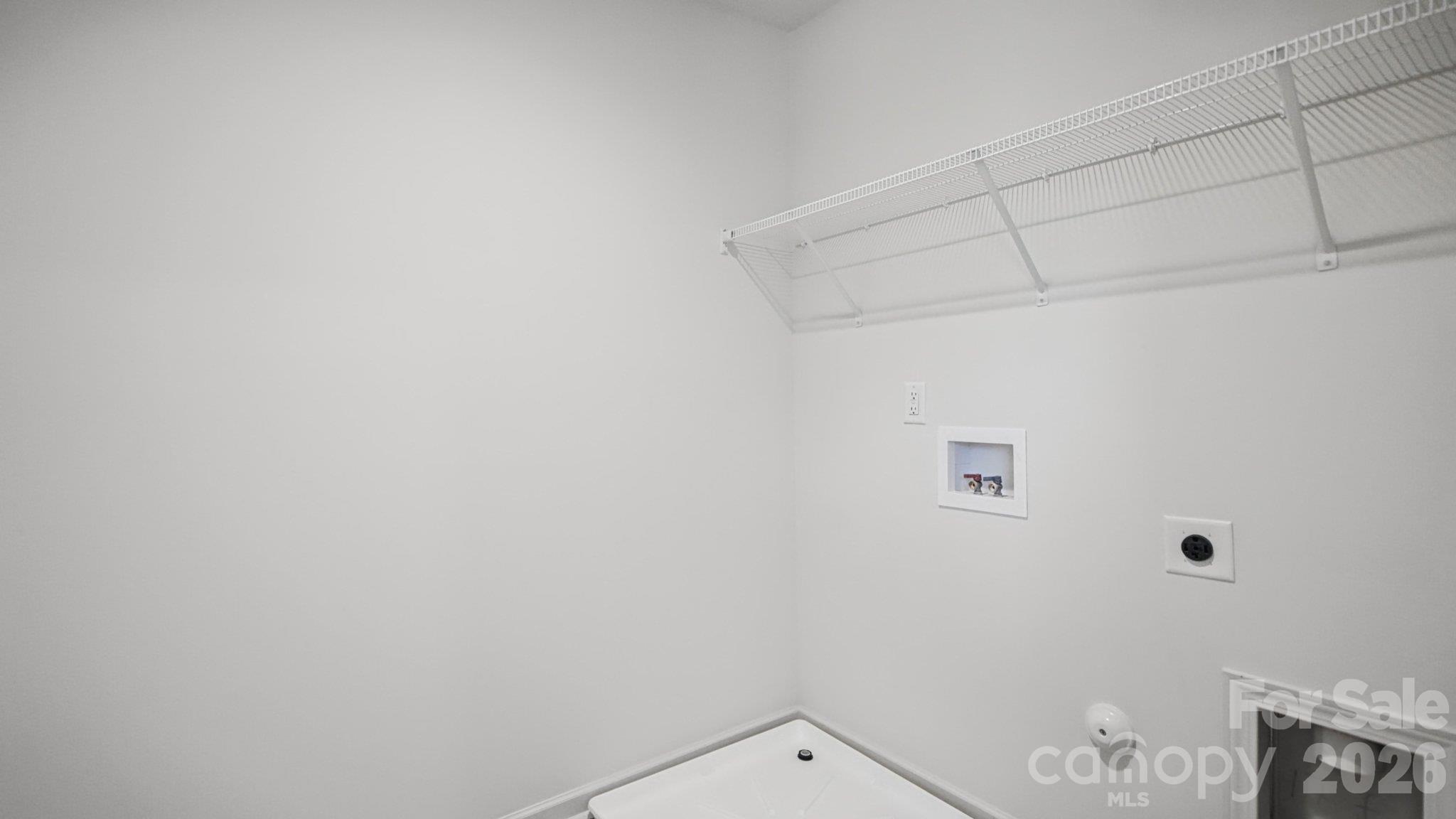 Laundry area with wire shelving, washer hookups, dryer outlet, and the edge of a white appliance.