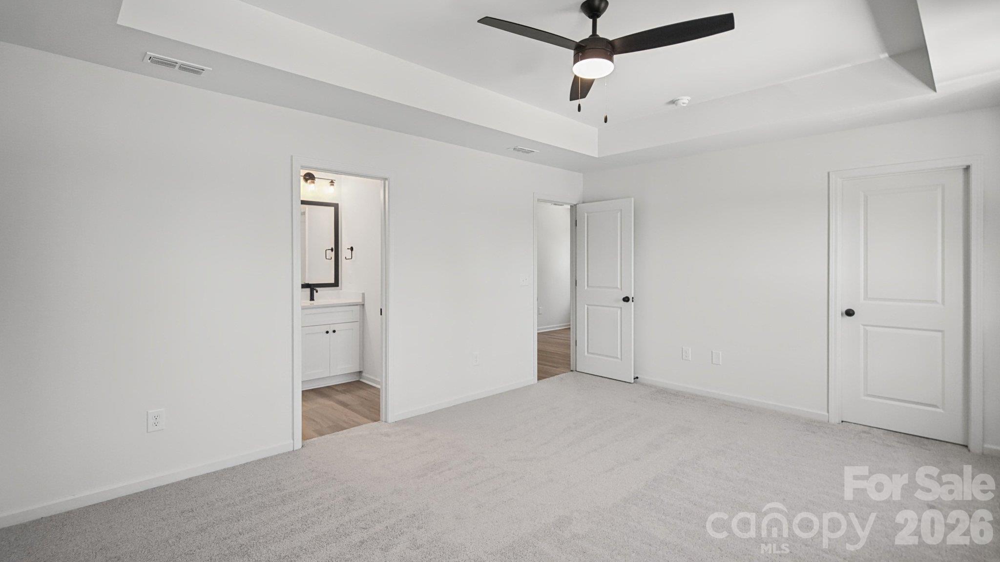 Empty carpeted bedroom with a tray ceiling and ceiling fan; open doorway reveals a bathroom vanity.