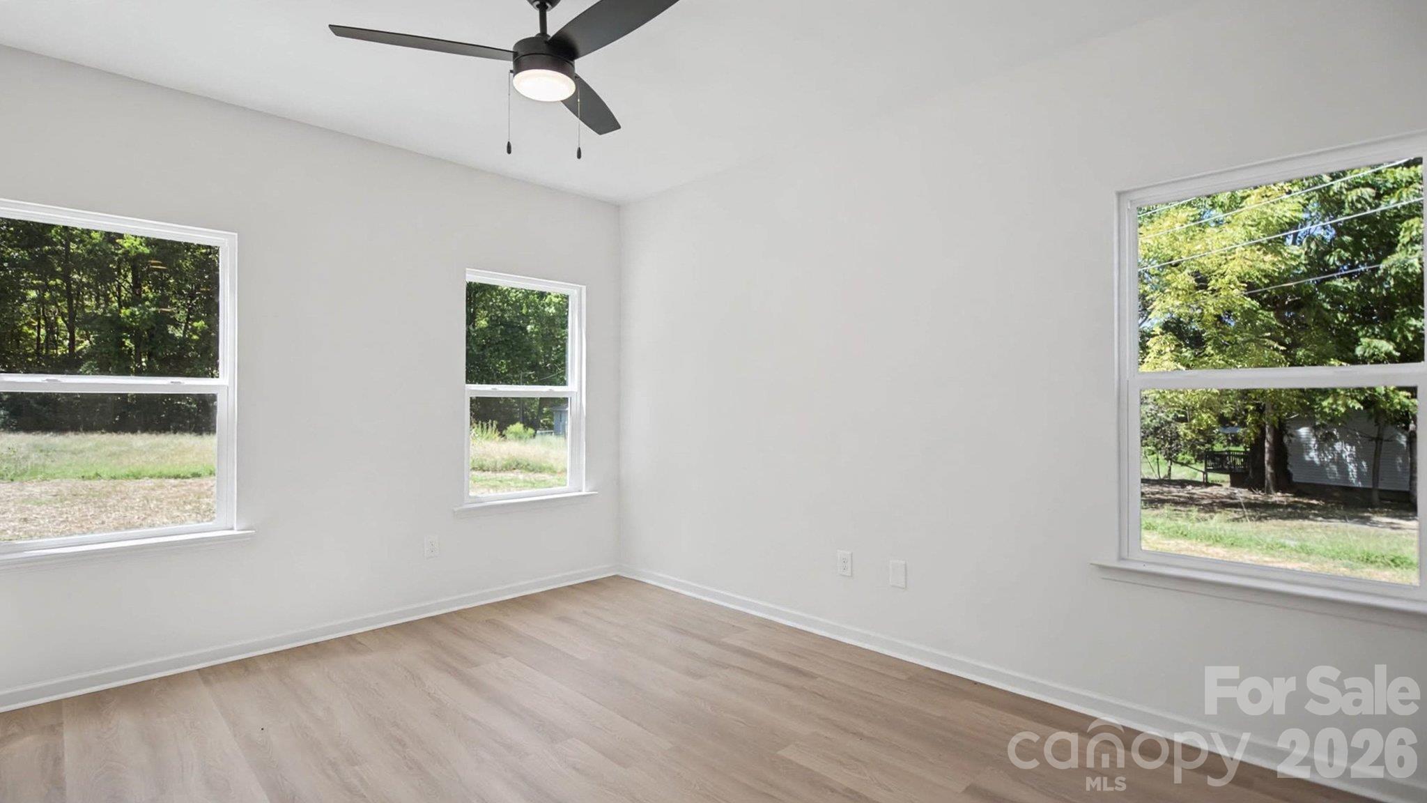 Vacant room with light wood-look floors, three large windows, and a black ceiling fan.