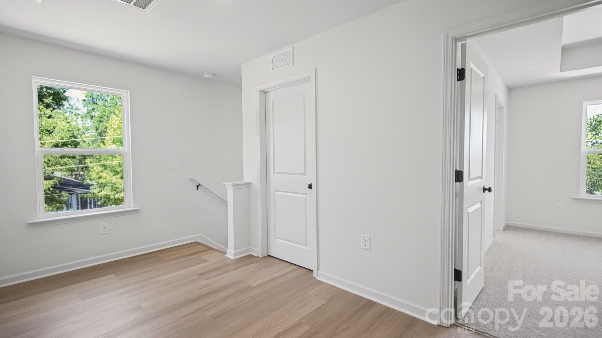 Empty bright room with light wood flooring, a window overlooking trees, and an open doorway to a carpeted room.