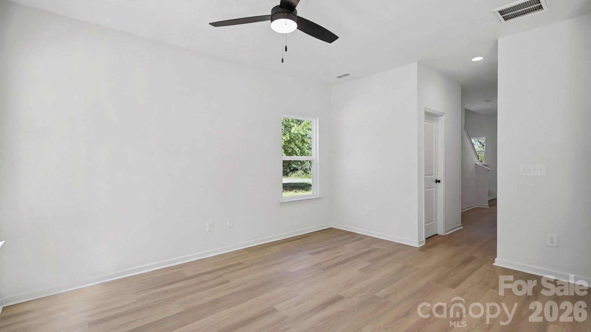 Vacant room with light wood floors, a black ceiling fan, and a window overlooking greenery.