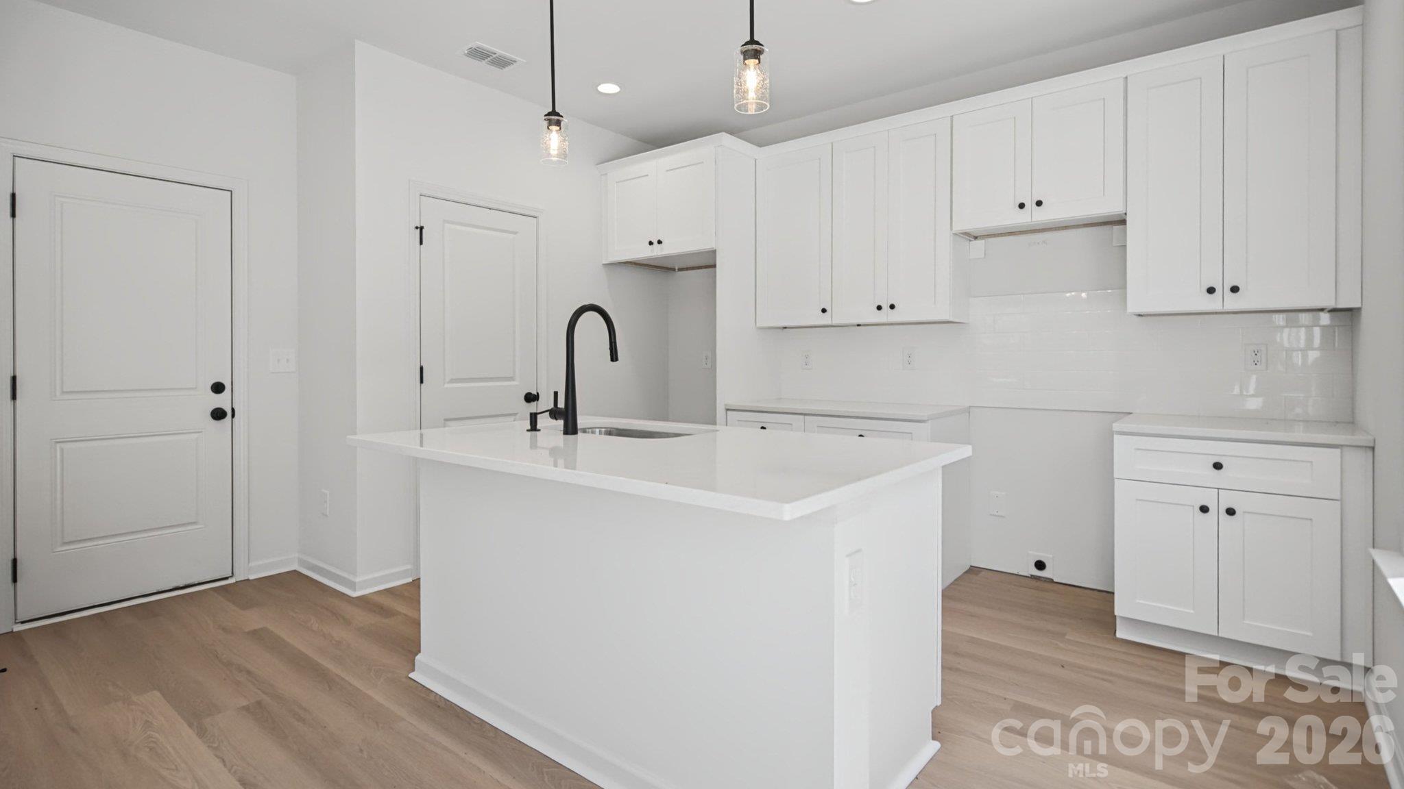 Bright white kitchen with a large island, black faucet, pendant lights, and light wood flooring.